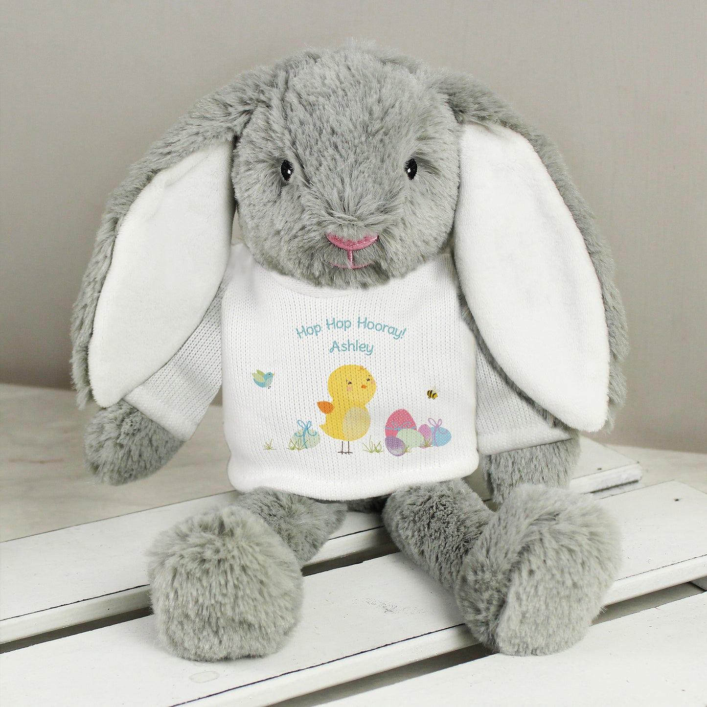Grey plush bunny with a white shirt featuring text and graphics on a wooden surface.