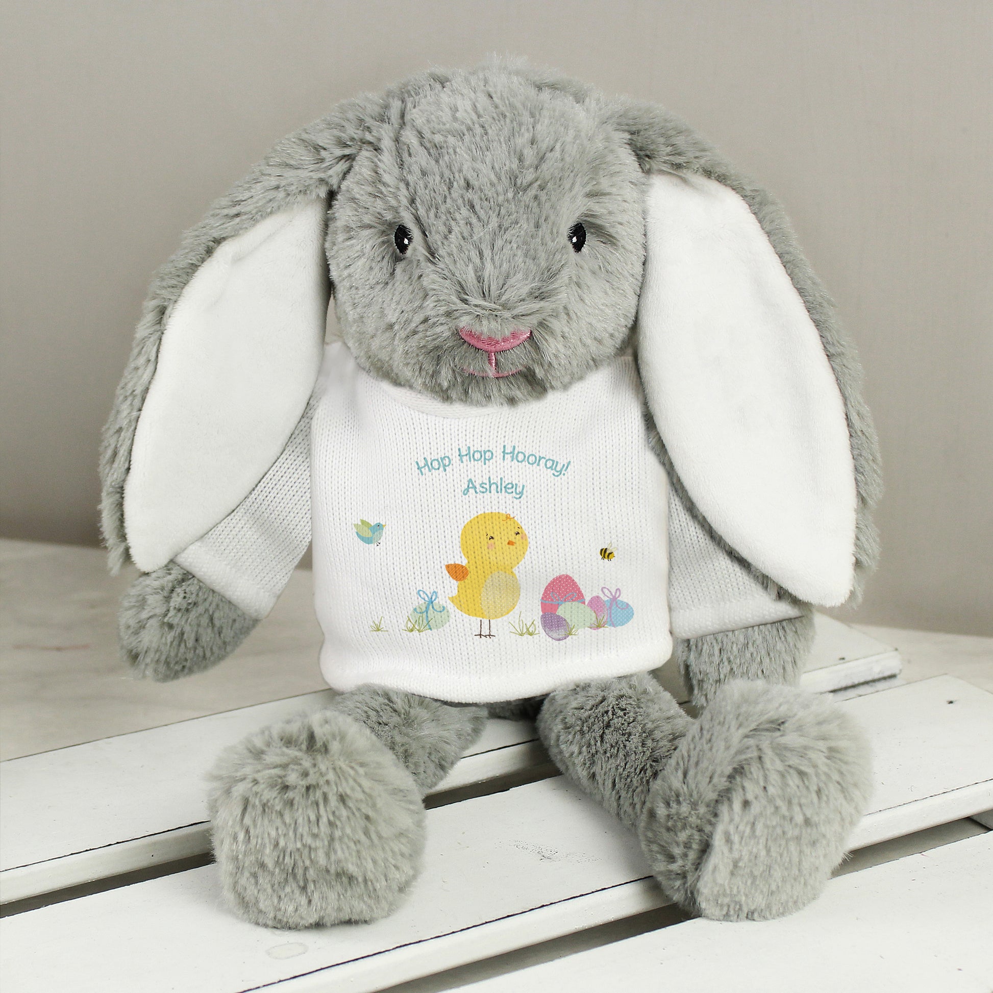 Grey plush bunny with a white shirt featuring text and graphics on a wooden surface.