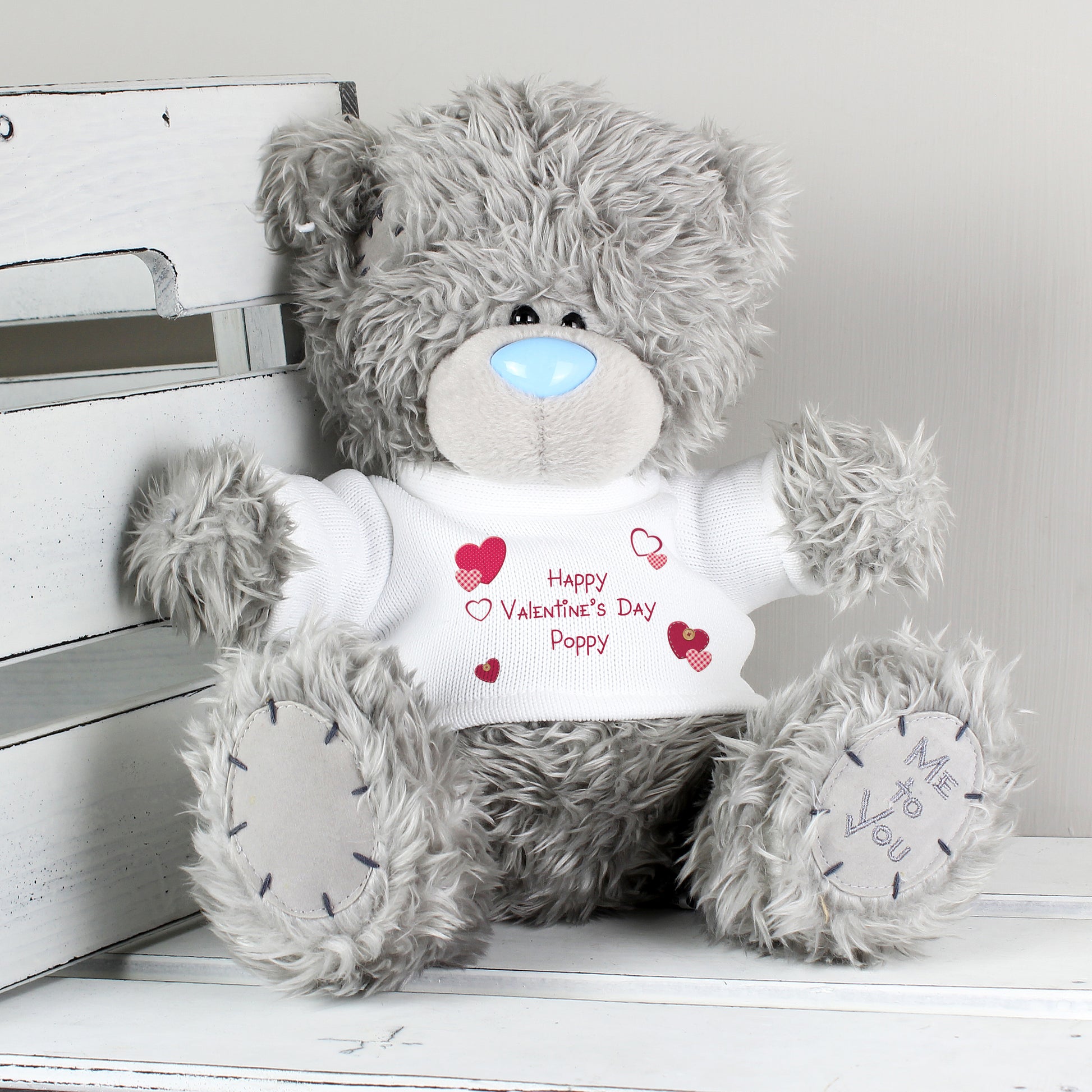 Grey teddy bear wearing a Valentine's Day shirt on a white wooden surface.