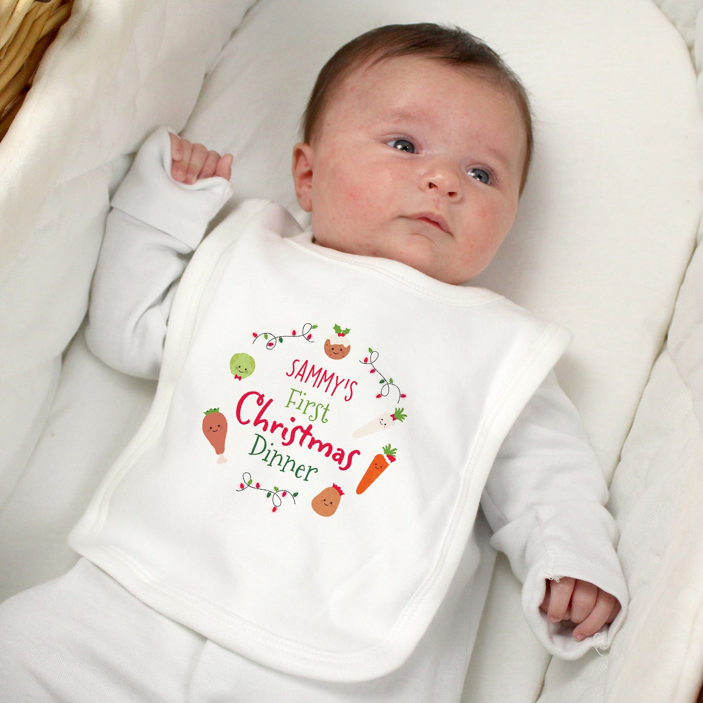 Baby wearing a bib with 'Sammy's First Christmas Dinner' text, lying on a white blanket.