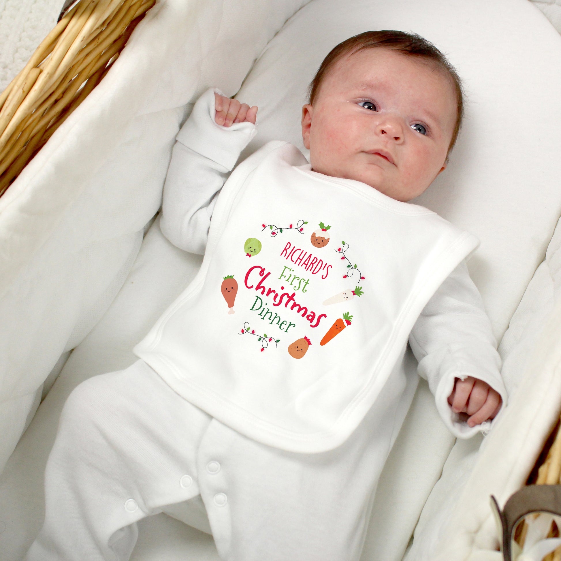 Baby wearing a personalised white onesie with 'Richard's First Christmas Dinner' text, lying on a white blanket.
