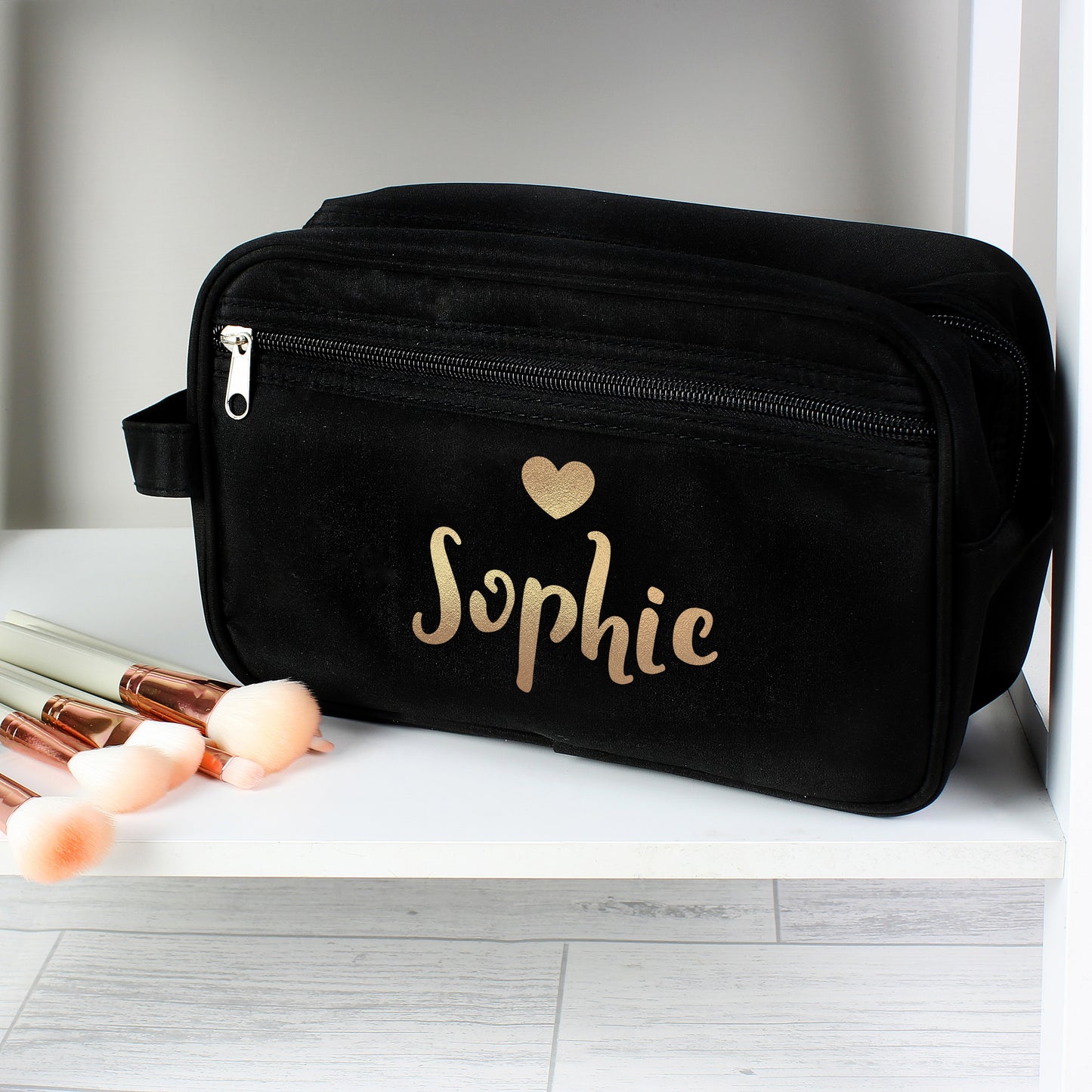 Black toiletry bag with 'Sophie' and a heart design on a white surface with makeup brushes.