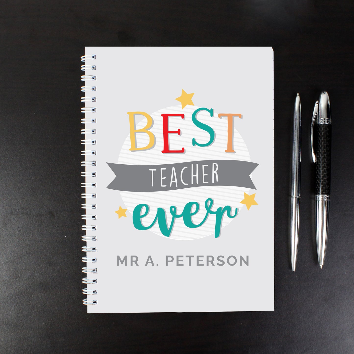 Notebook with 'Best Teacher Ever' text and two pens on a dark background