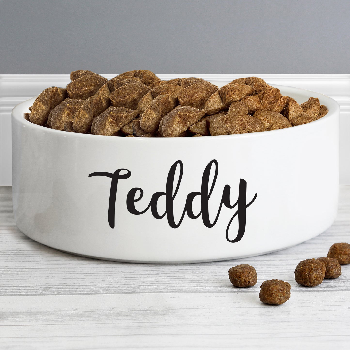White pet bowl with 'Teddy' on it, filled with dog food, on a wooden surface.