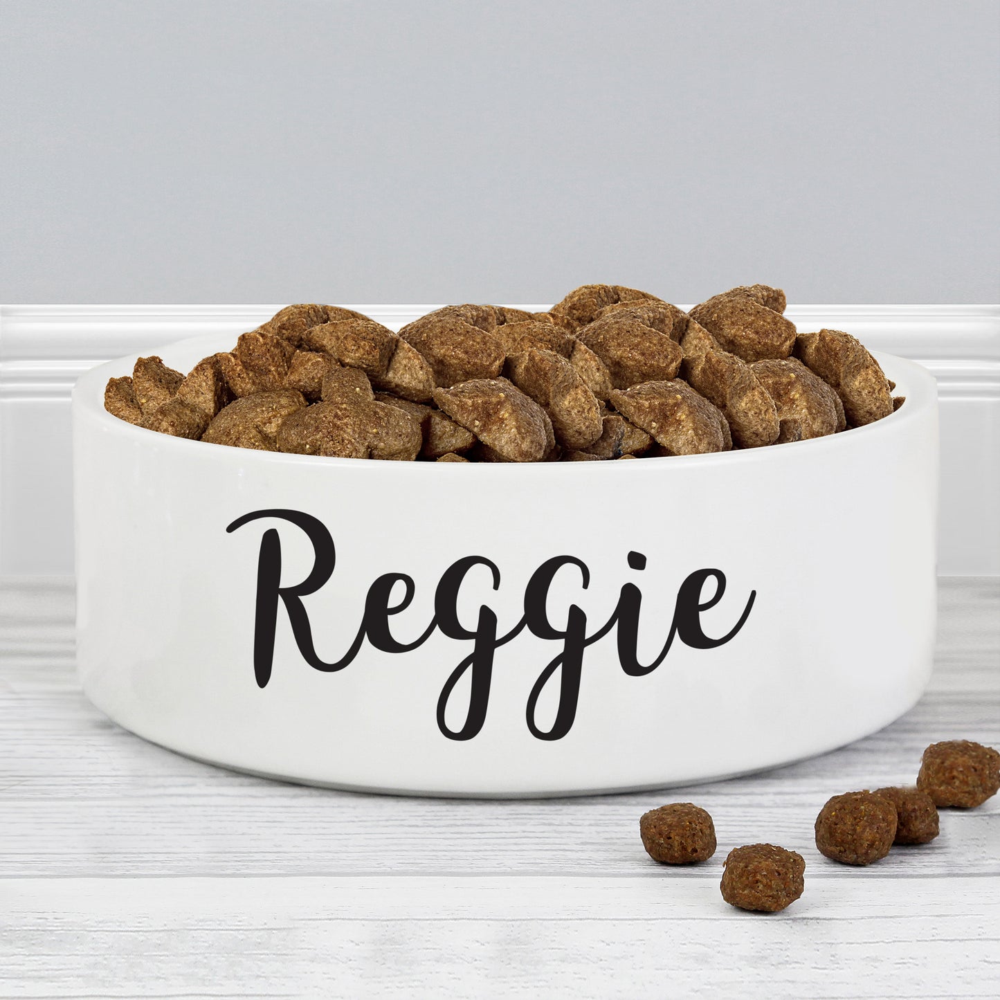 Personalised White pet bowl labeled 'Reggie' with dry dog food on a light gray background