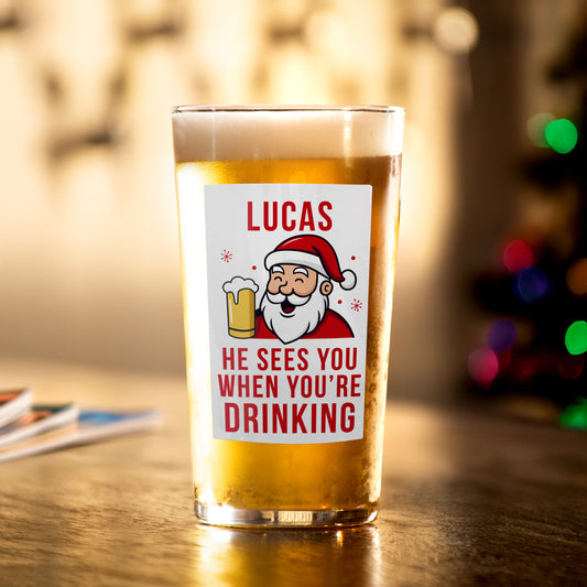 Personalised Beer glass with Santa Claus label and text 'Lucas sees you when you're drinking' on a wooden table.