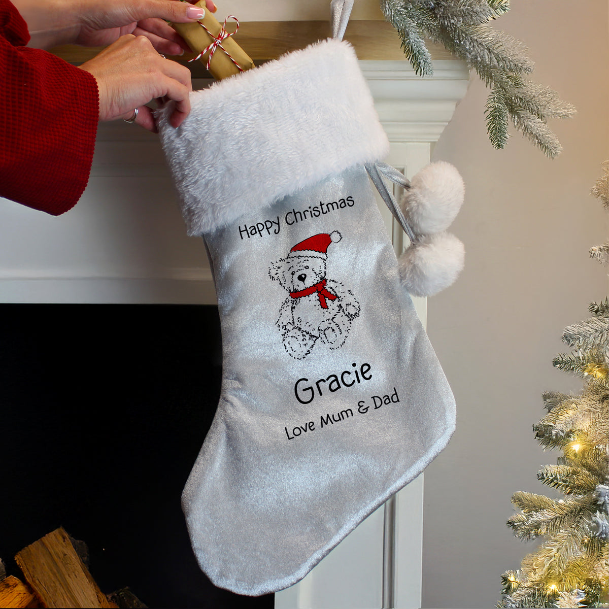 Grey Christmas stocking with a teddy bear and text, held by a person against a festive background.