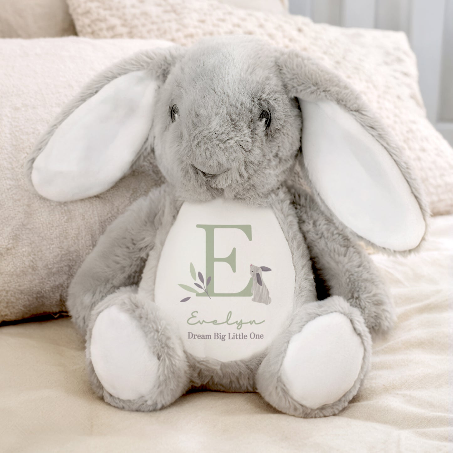 Grey plush bunny with a white belly featuring the letter 'E' and text, sitting on a soft surface.