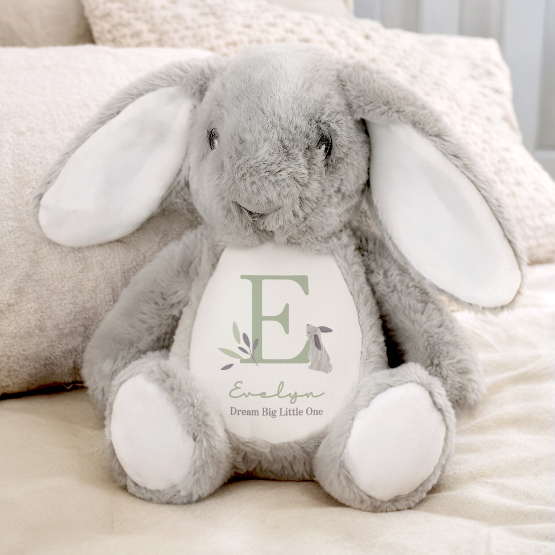 Grey plush bunny with a white belly featuring the letter 'E' and text, sitting on a soft surface.
