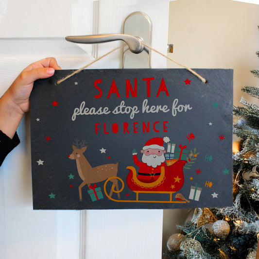 Hand holding a Personalised Christmas sign with Santa and reindeer, text 'Santa please stop here for Florence', against a white door and decorated tree background.