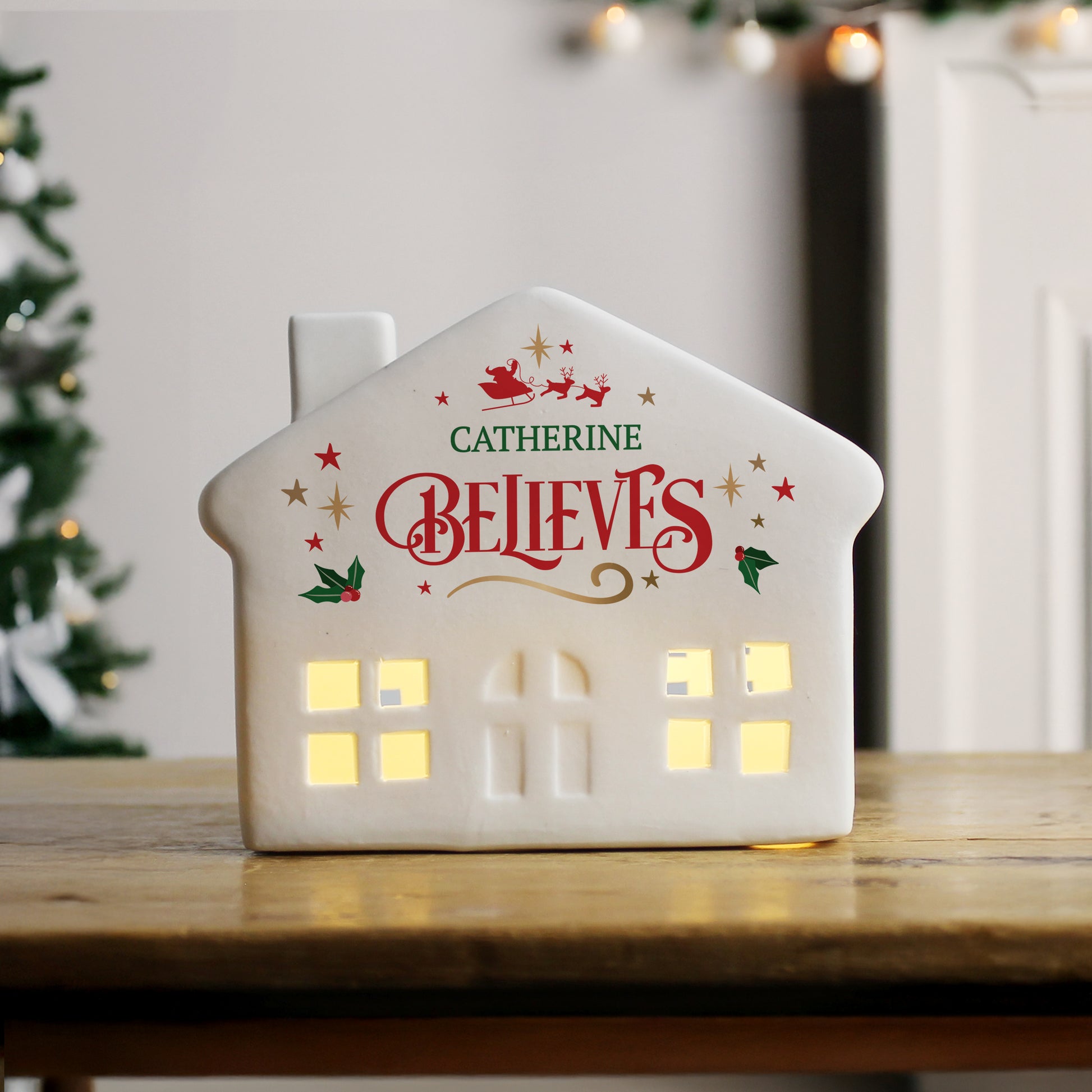 Decorative house-shaped light with 'Catherine Believes' text on a wooden surface with a Christmas tree in the background.