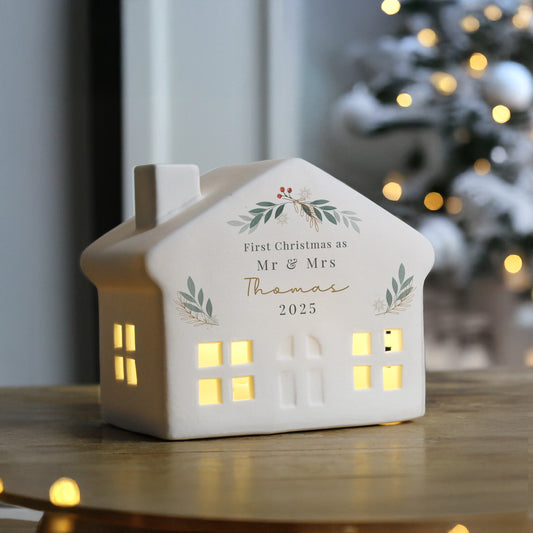 Decorative house-shaped lantern with personalised text on a table, blurred Christmas tree in the background.