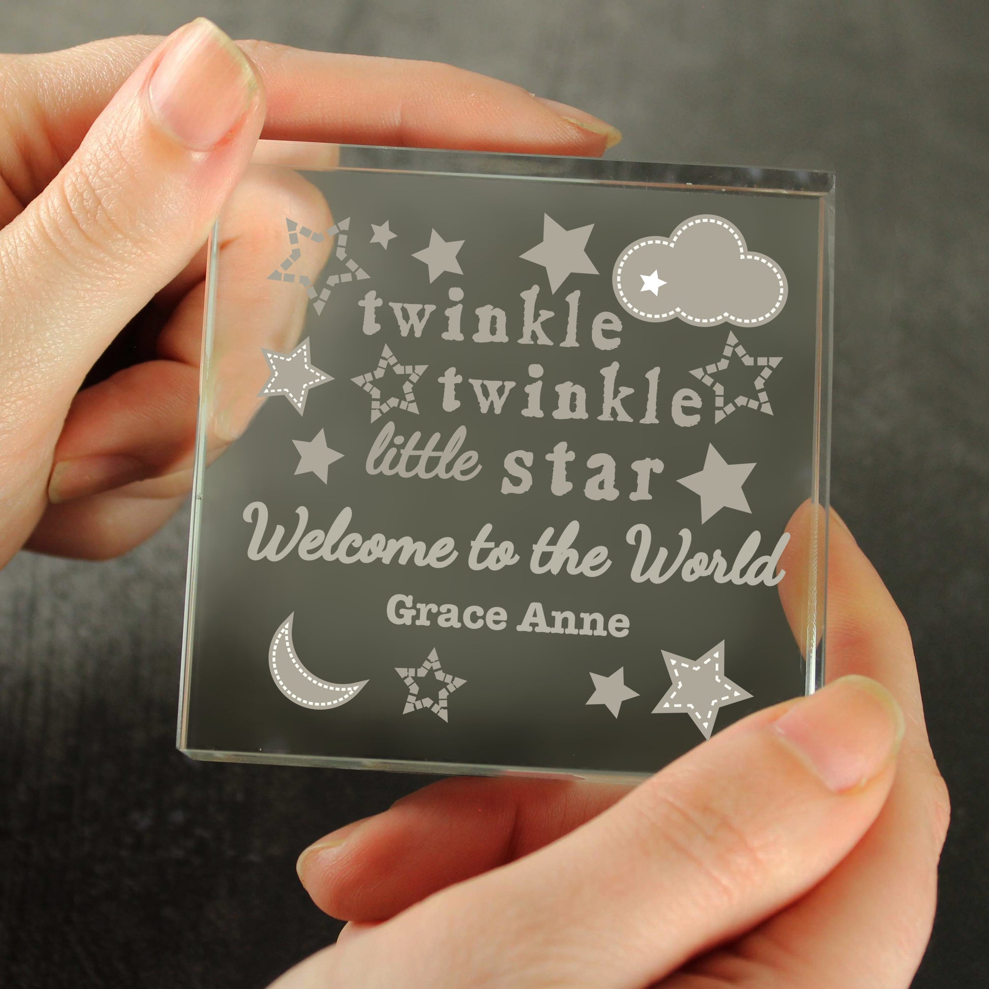 Clear glass block with engraved text 'twinkle twinkle little star Welcome to the World Grace Anne' held by a hand against a dark background.