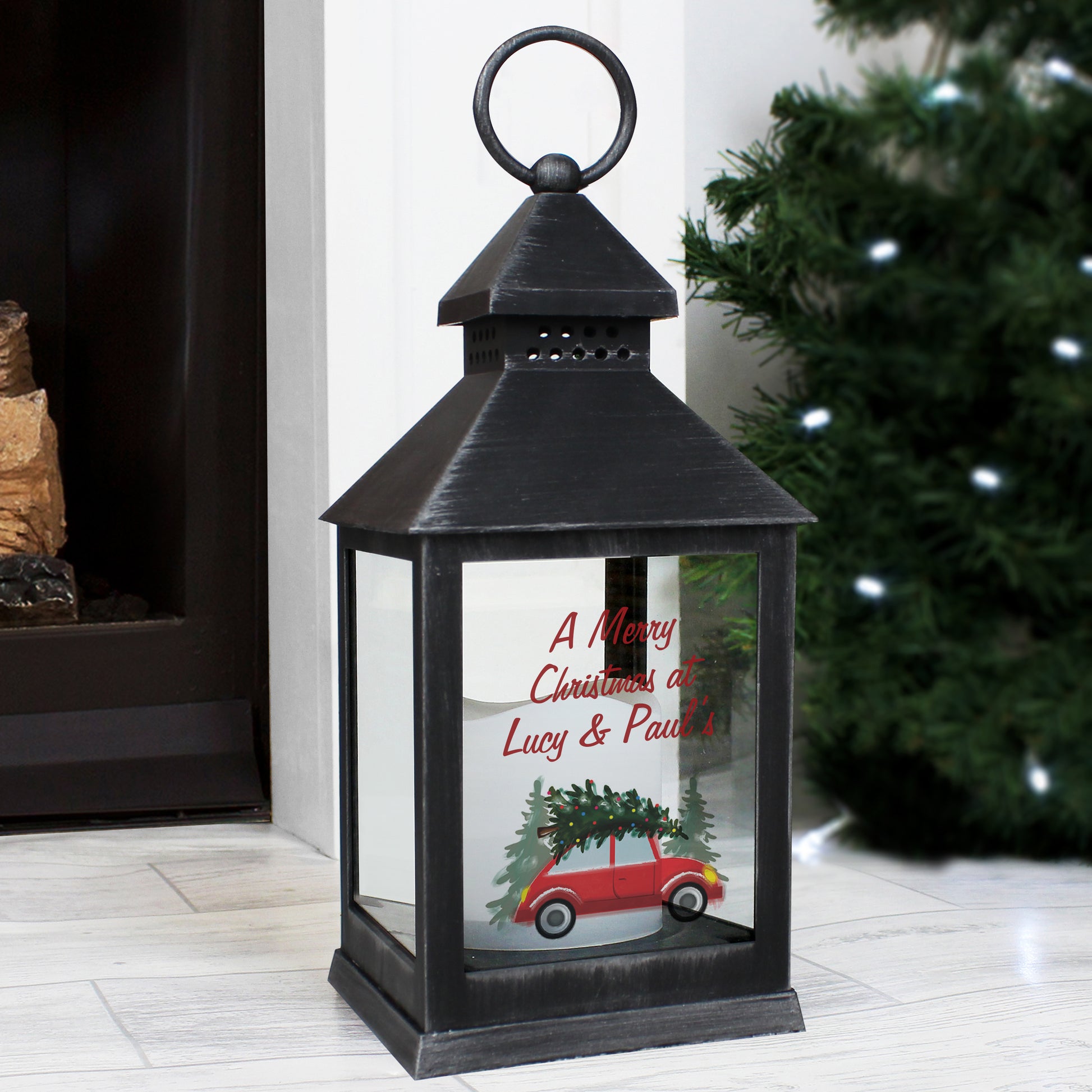 Black lantern with Christmas design and personalised text in front of a fireplace and tree.