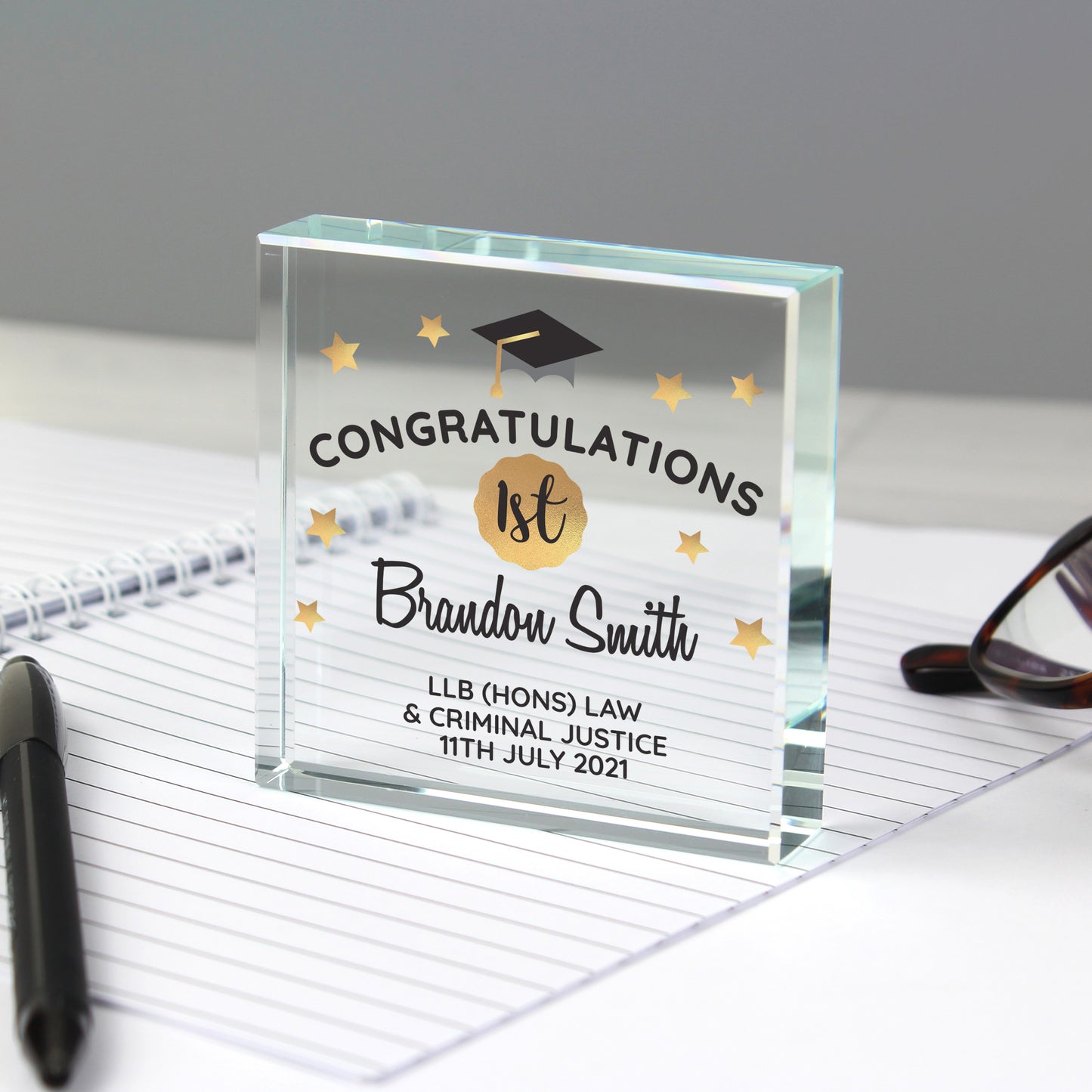 Glass graduation award with personalised text on a desk with glasses and pen