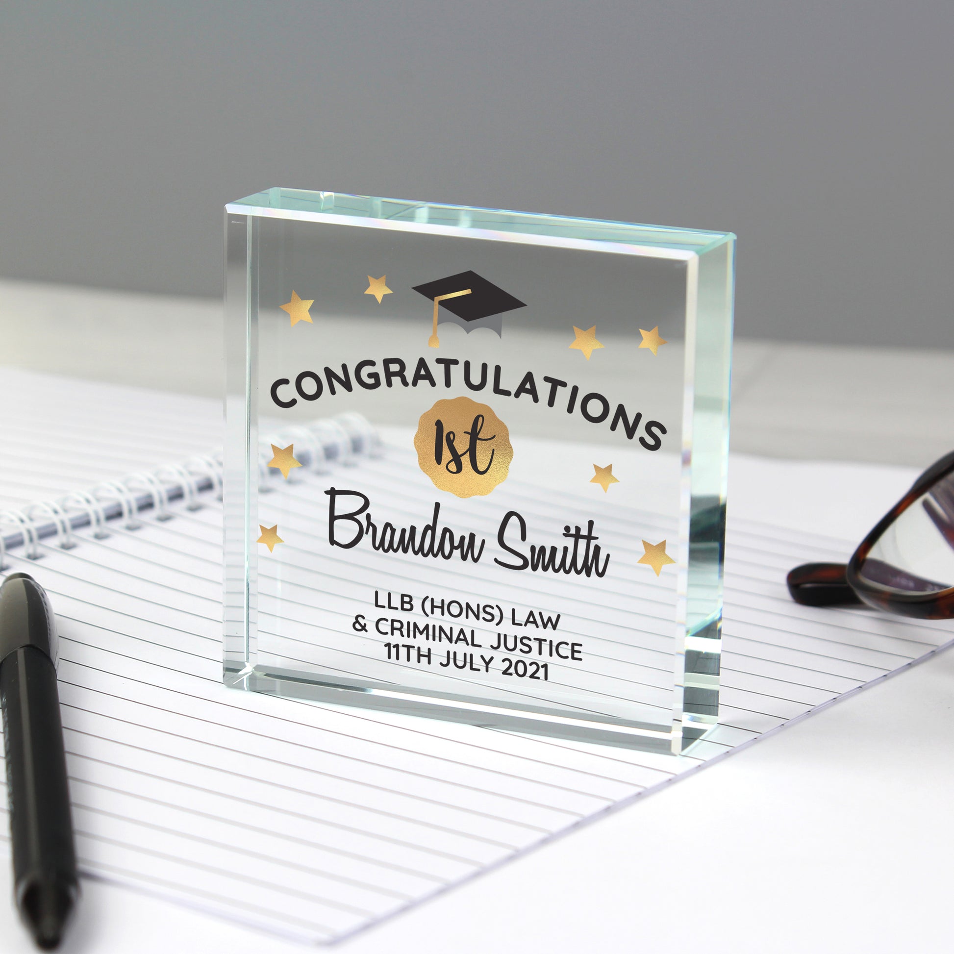 Glass graduation award with personalised text on a desk with glasses and pen