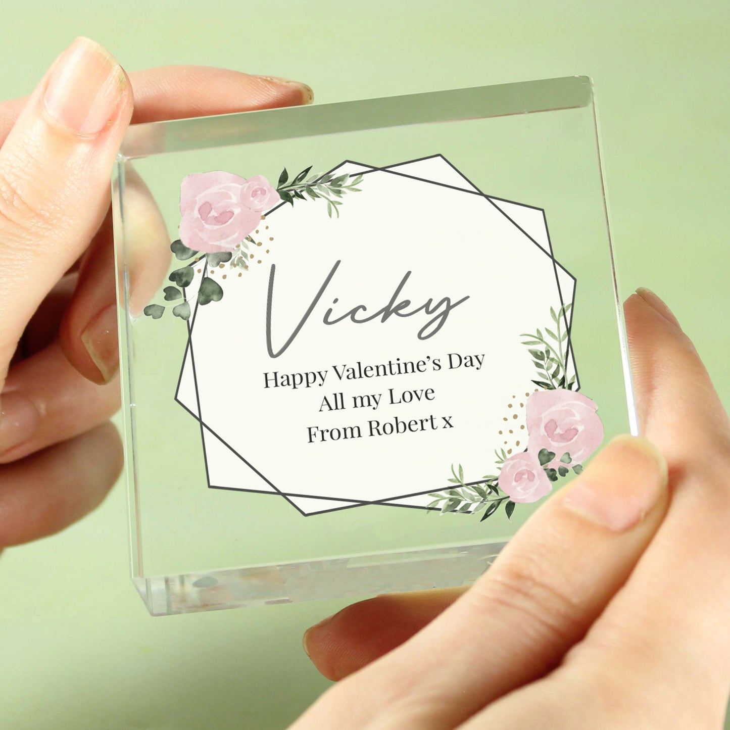 Clear glass block with floral design and personalized message held by hands against a light green background