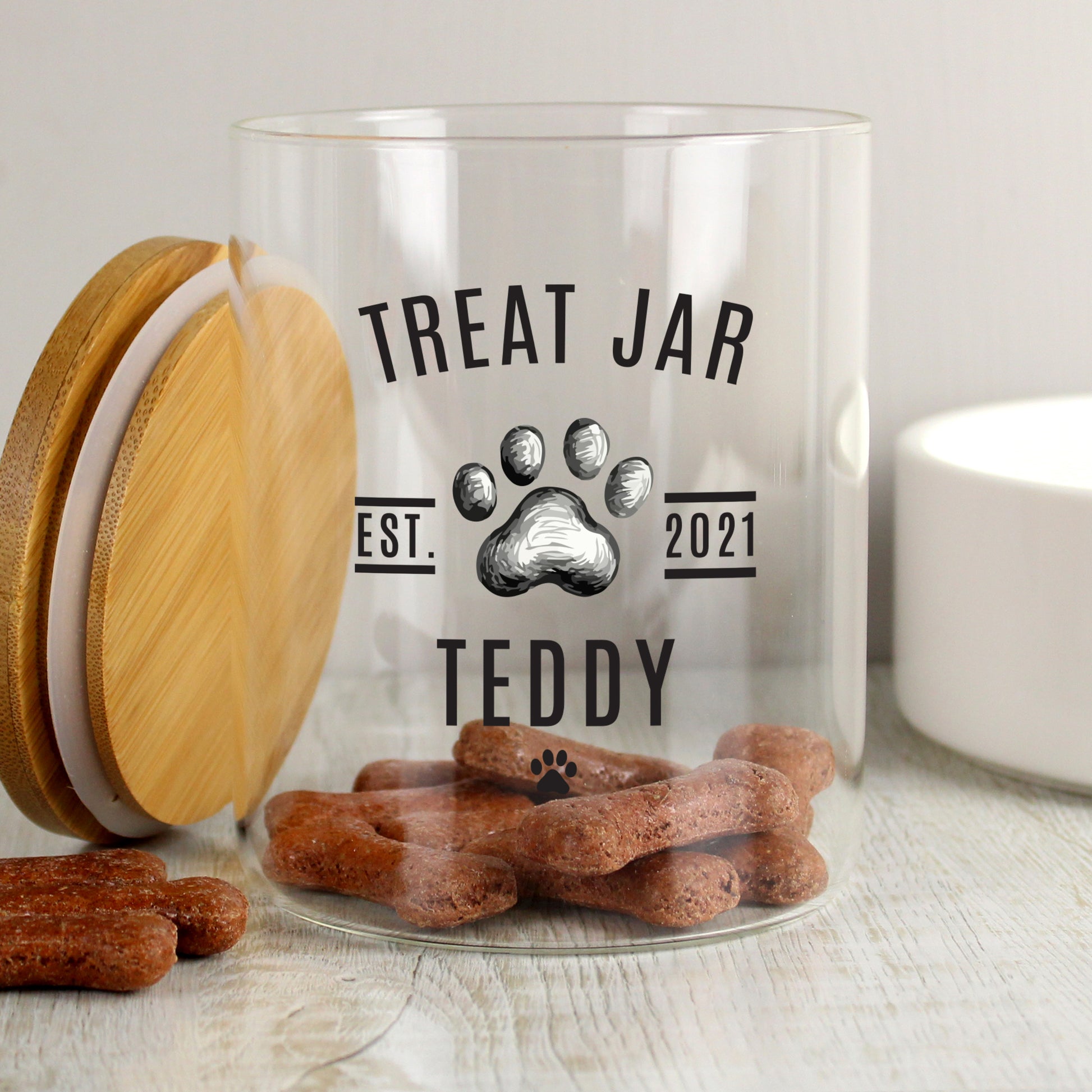Clear treat jar with wooden lid labeled 'Treat Jar Teddy' filled with dog treats on a light surface.