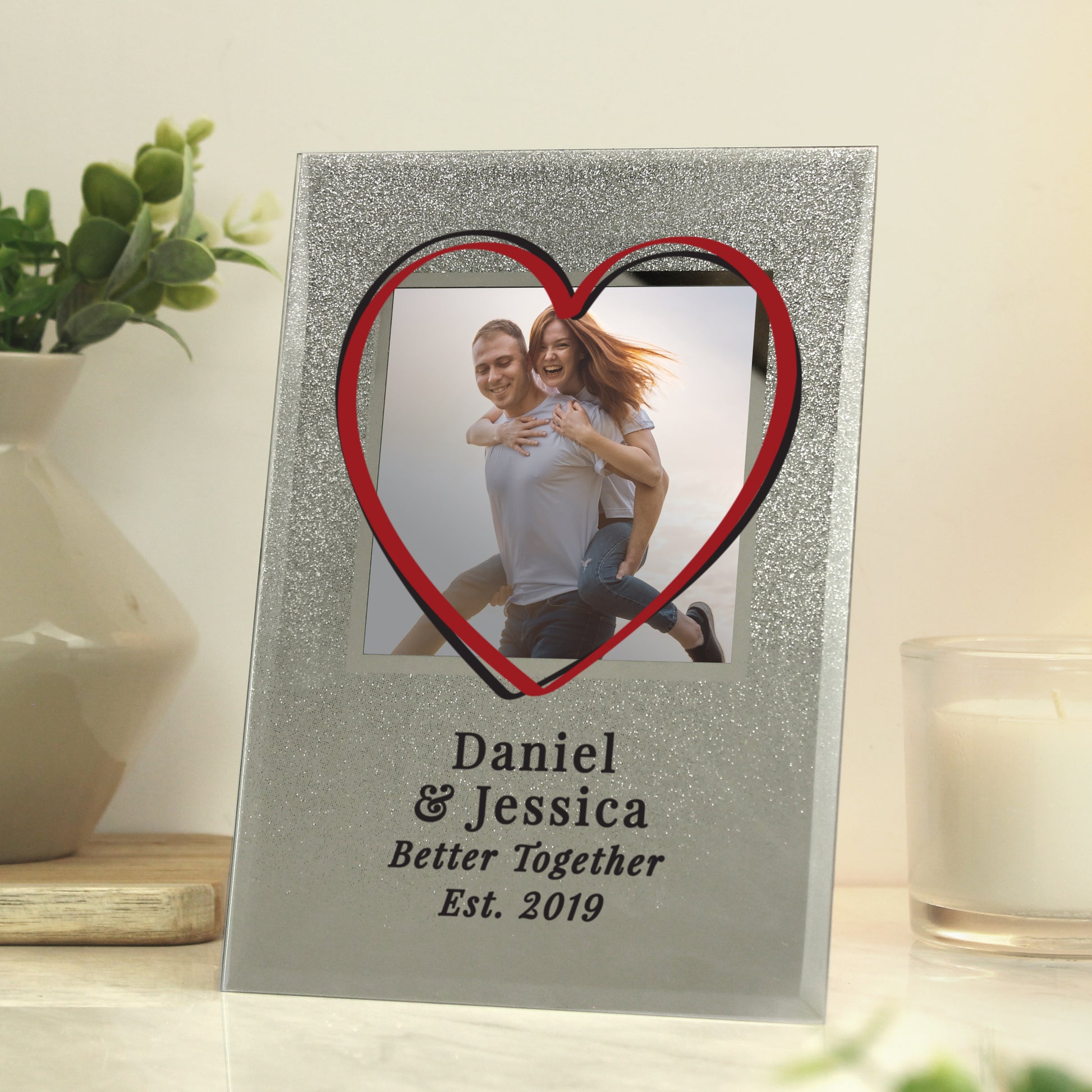 Personalised photo frame with heart design displaying a couple, placed on a surface with a plant and candle in the background.