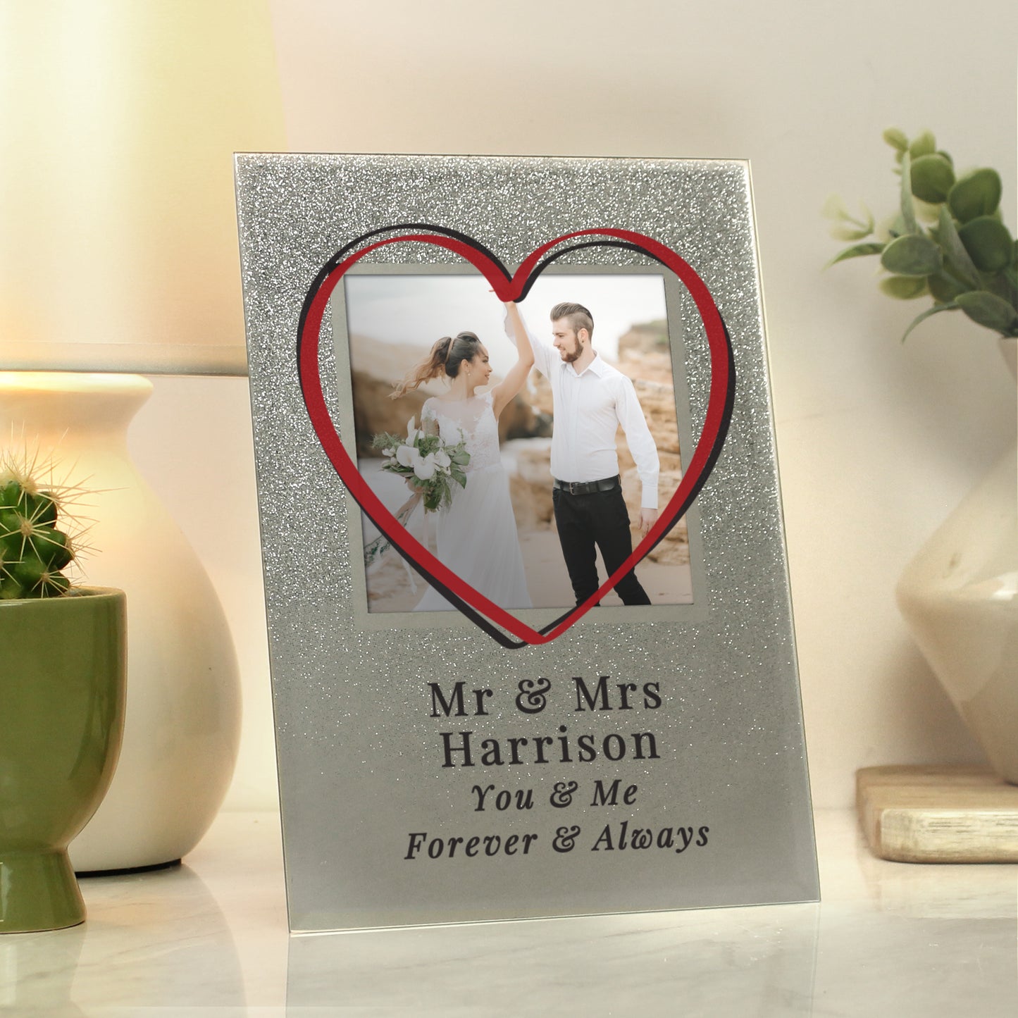 Personalised photo frame with a heart-shaped cutout and engraved text, placed on a surface with decorative items.
