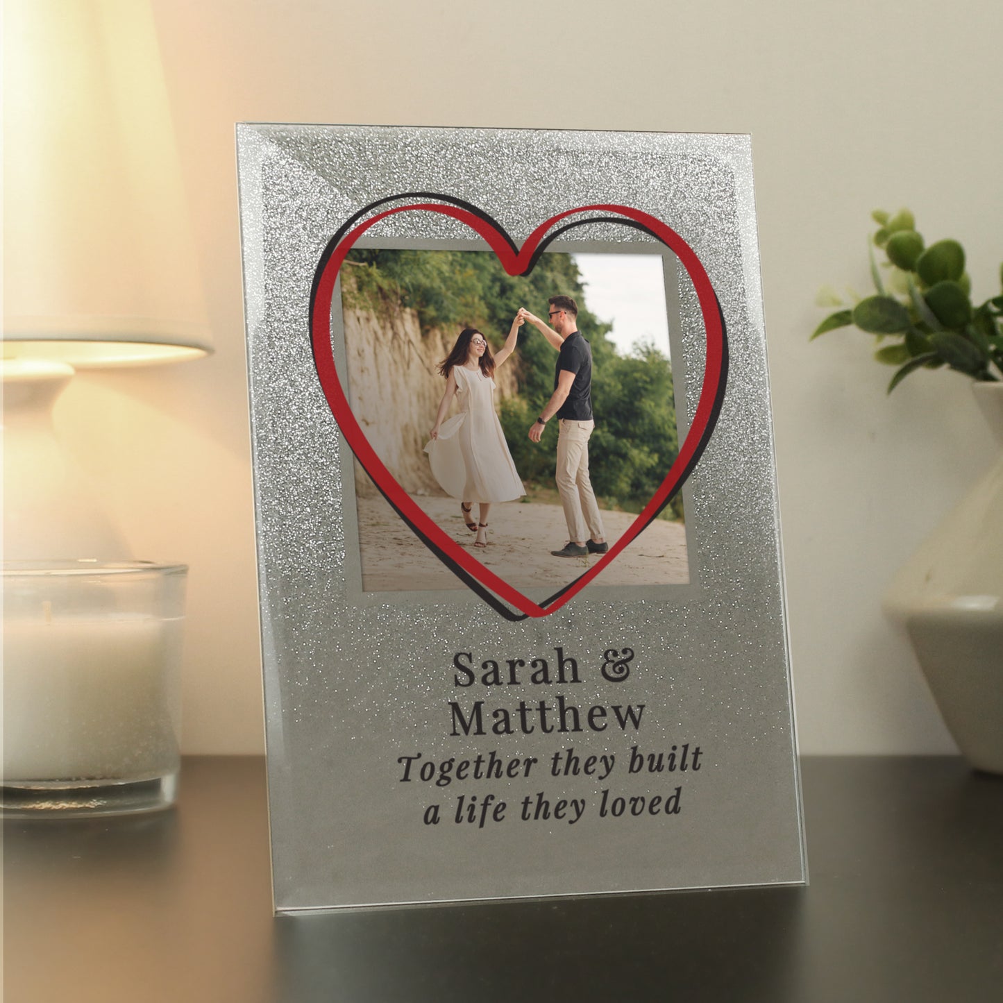 Decorative photo frame with a heart-shaped cutout and couple's name and quote on a table.