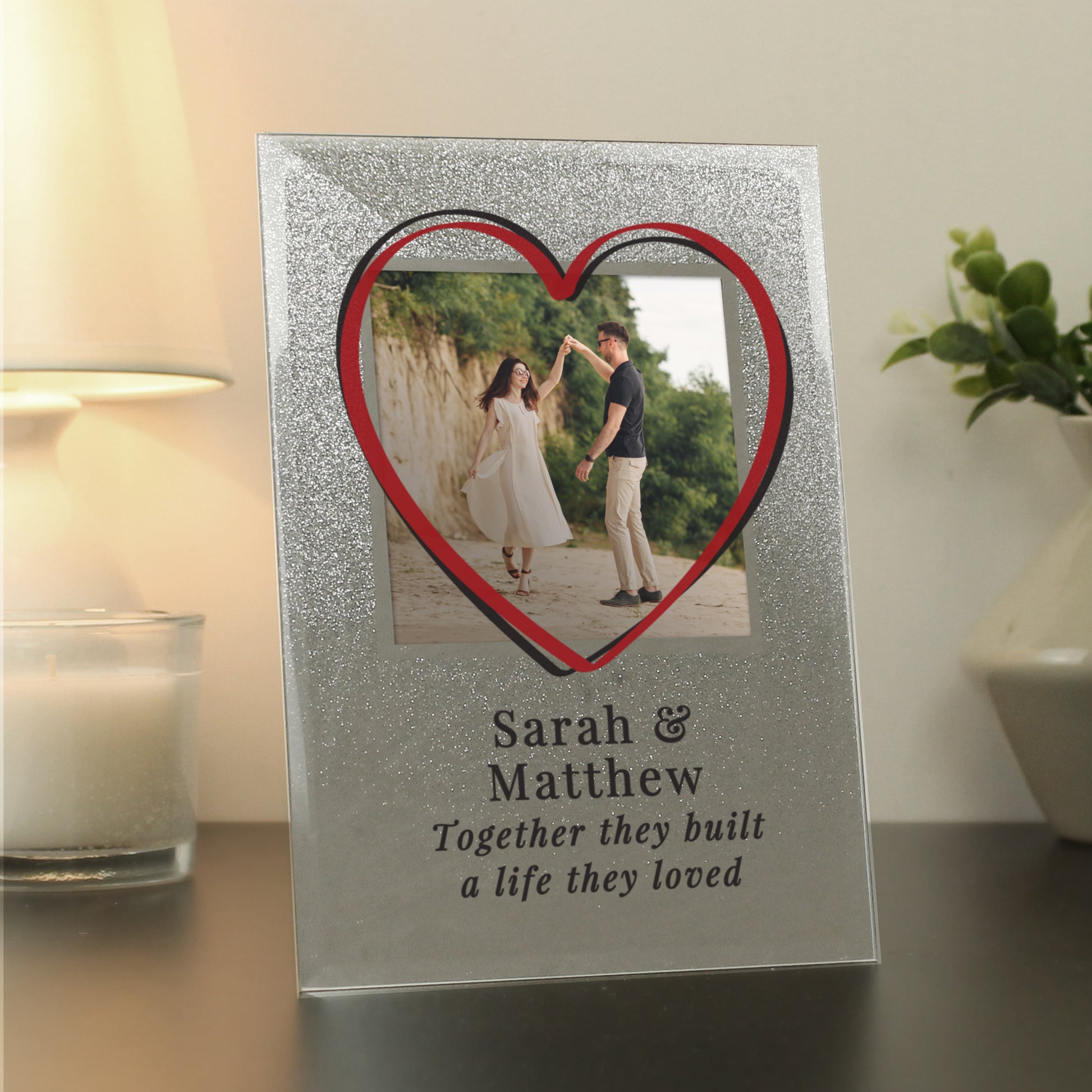 Decorative photo frame with a heart-shaped cutout and couple's name and quote on a table.