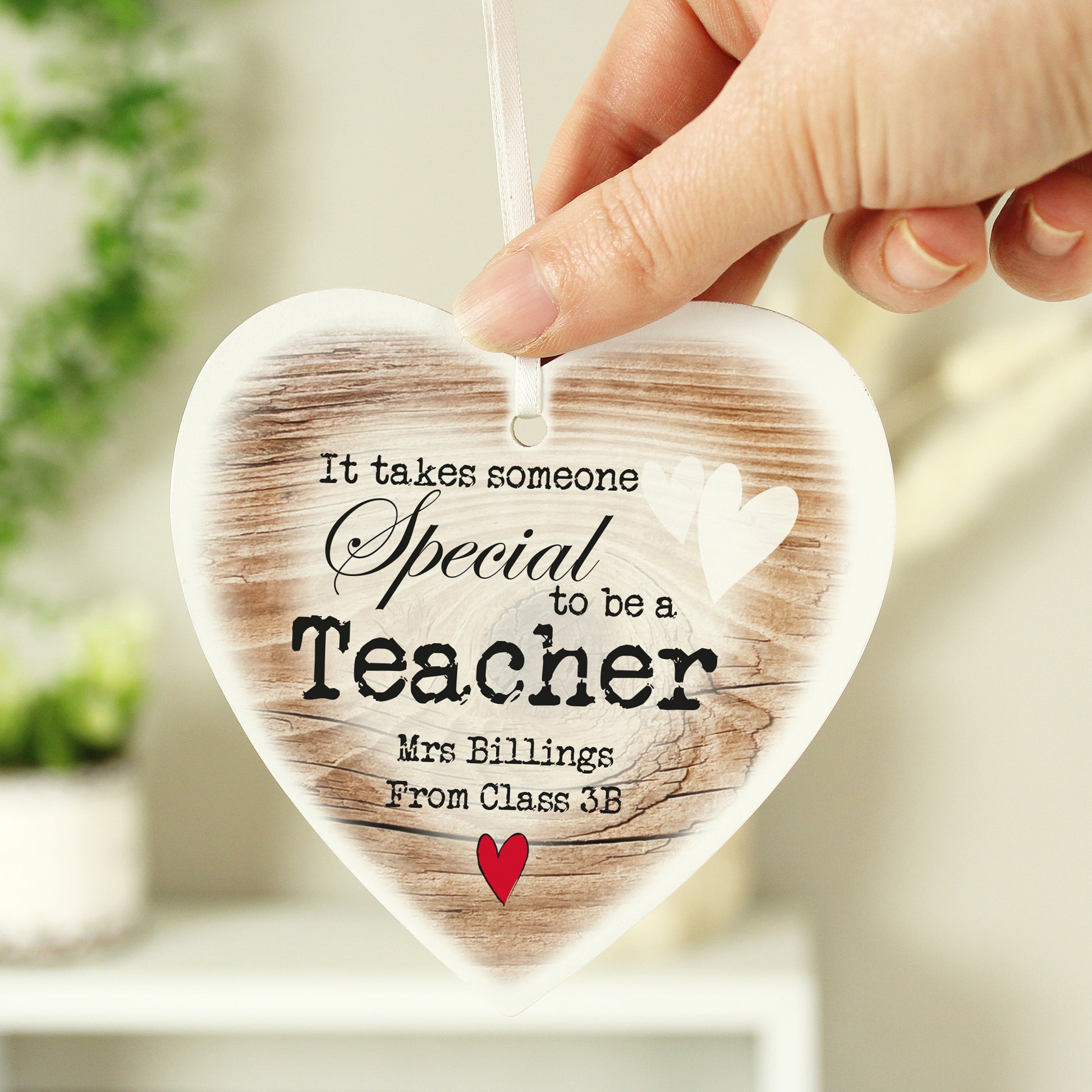 Heart-shaped wooden ornament with printed text, held by a hand against a blurred indoor background.