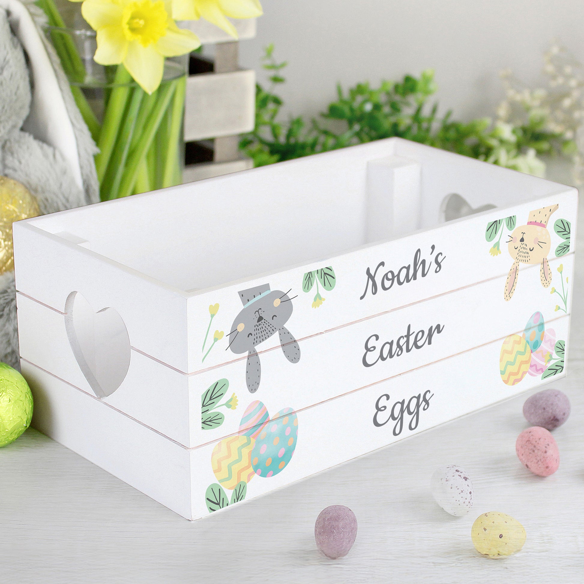 White Easter egg crate with decorative design and text, surrounded by Easter eggs and flowers.