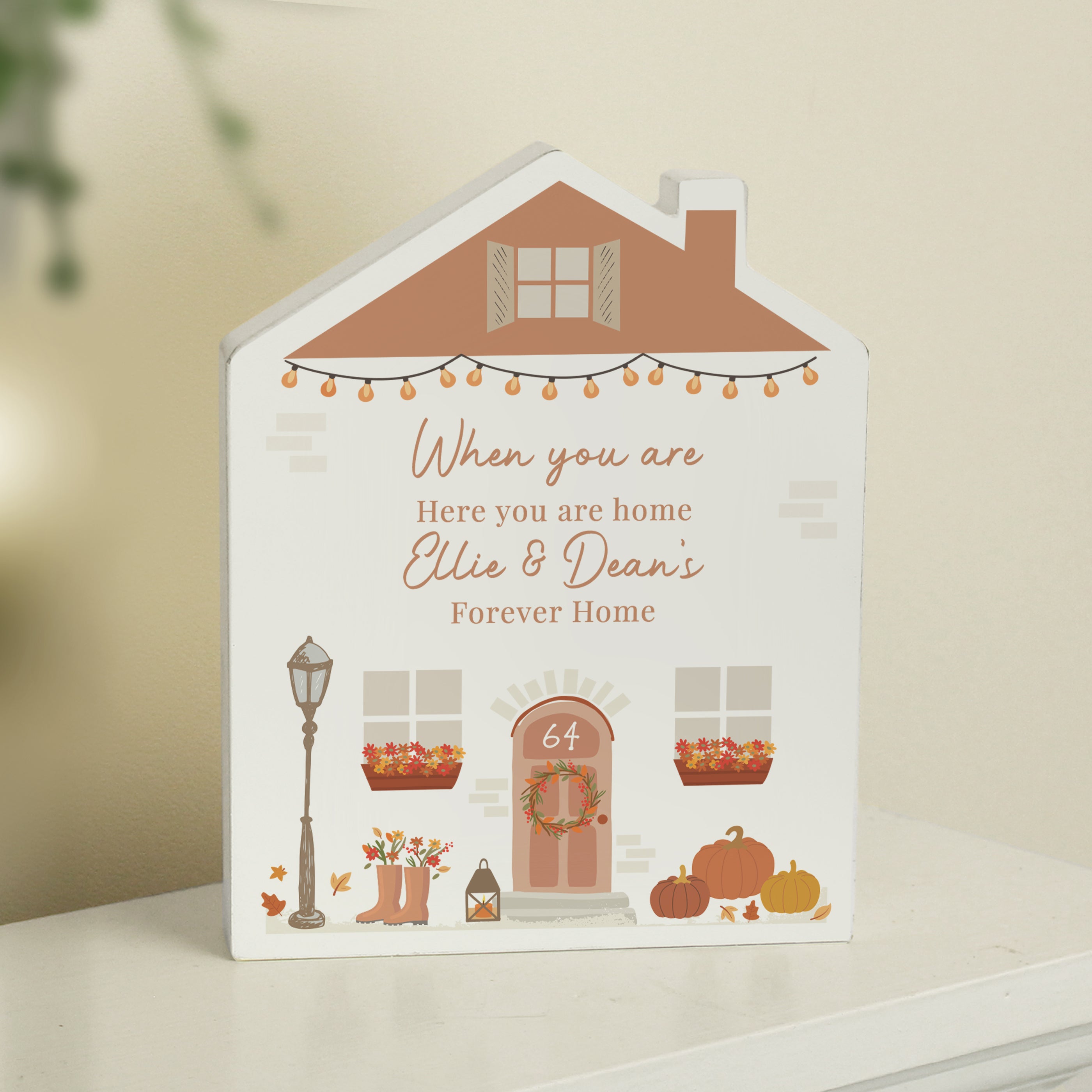 Personalised wooden house ornament with an autumn design, featuring a terracotta roof, pumpkin string lights, and a front door with the number "64" and a seasonal wreath. Decorative touches include pumpkins, a streetlamp, flower-filled boots, and planter boxes under the windows. The printed message includes placeholder names and a cosy quote about home, making it ideal for celebrating a new home or the autumn season with custom details.