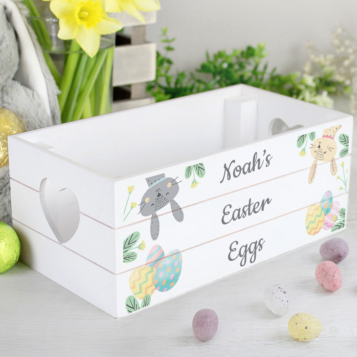 White Easter egg crate with decorative design and text, surrounded by Easter eggs and flowers.