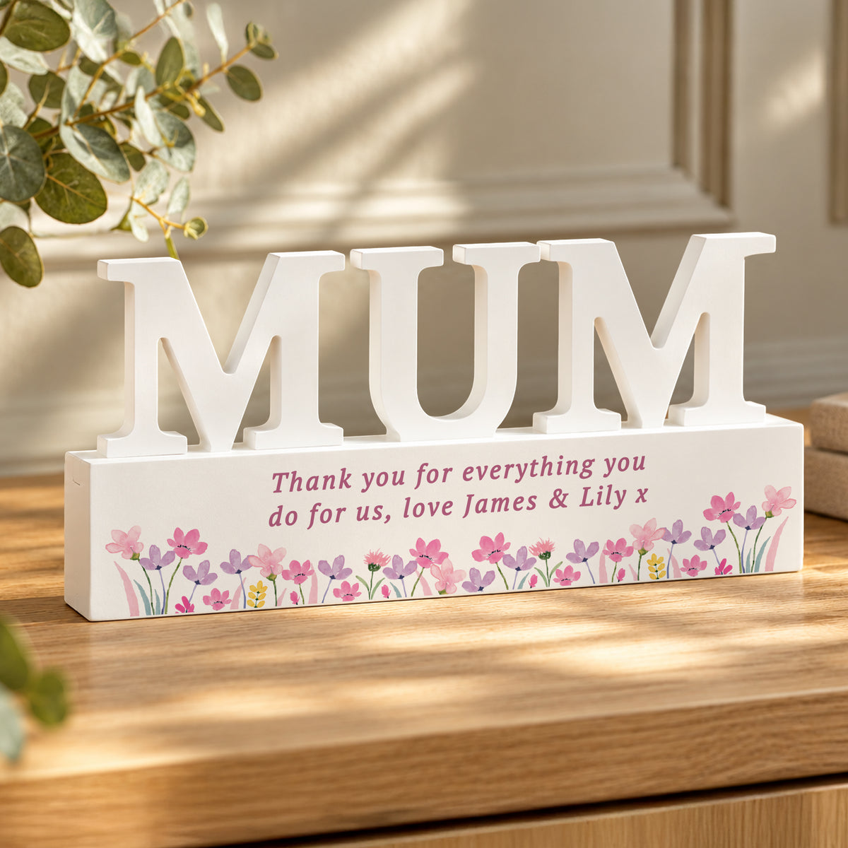 A white wooden decorative sign spelling “MUM” in large letters, mounted on a rectangular base printed with pink floral flowers and a personalised message in pink text. The sign sits on a wooden table with soft sunlight and blurred greenery in the background.