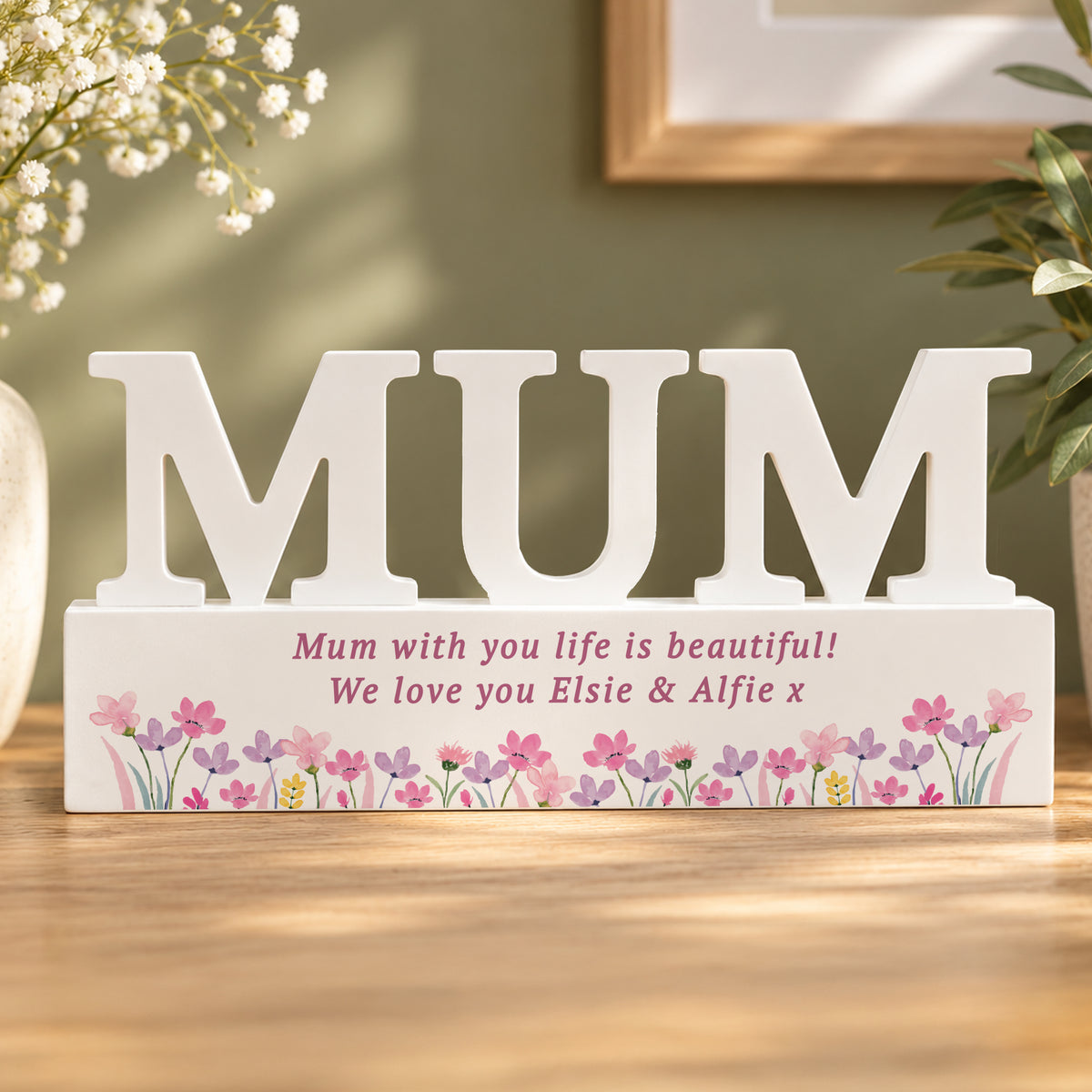 A white personalised wooden decorative sign spelling 'MUM' in large letters, placed on a tabletop. The base is printed with pink floral decoration and the message 'Mum with you life is beautiful! We love you Elsie & Alfie x' in pink text.