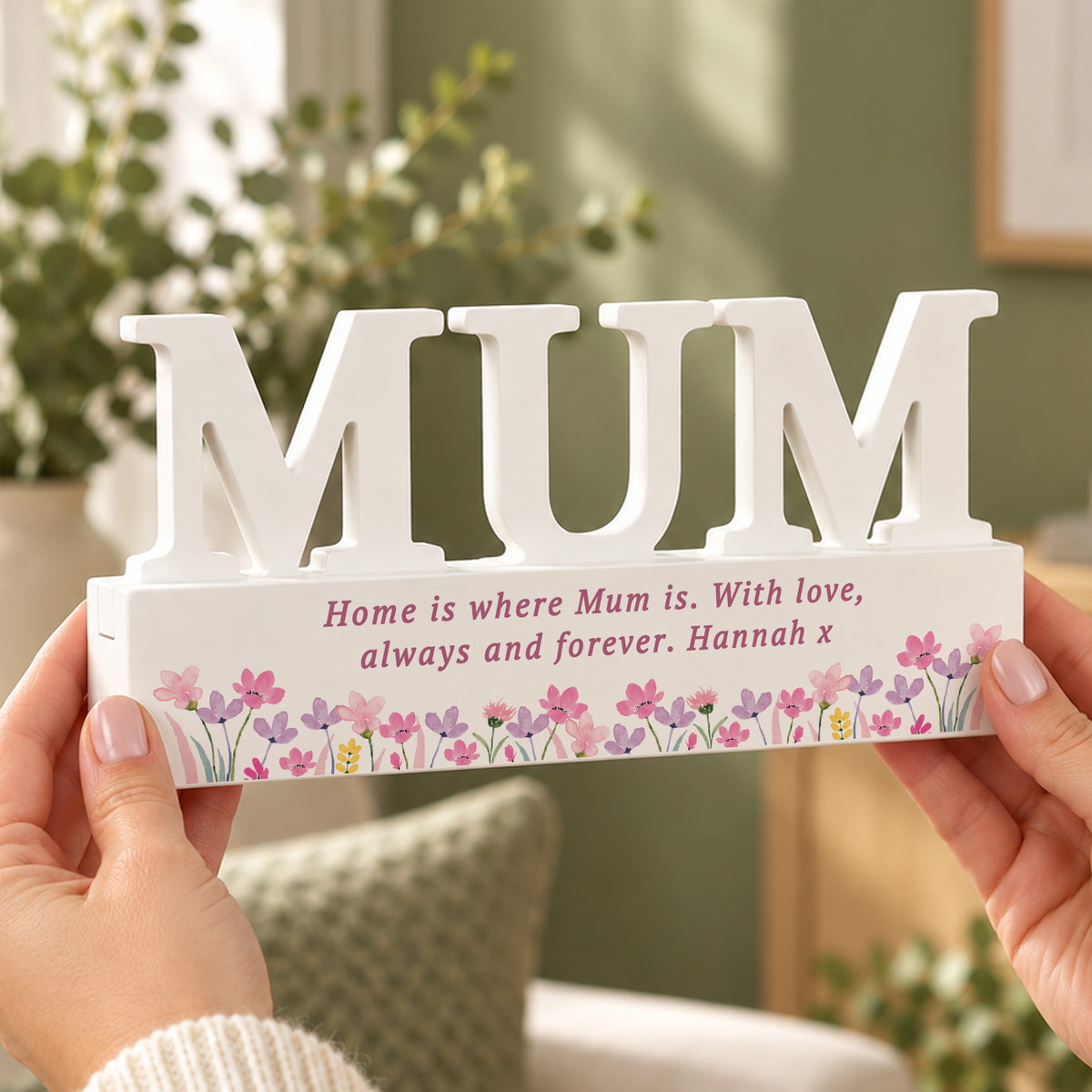 A person holds a white decorative wooden sign spelling “MUM” with a personalised message and floral border on the base, shown in a softly lit home interior.