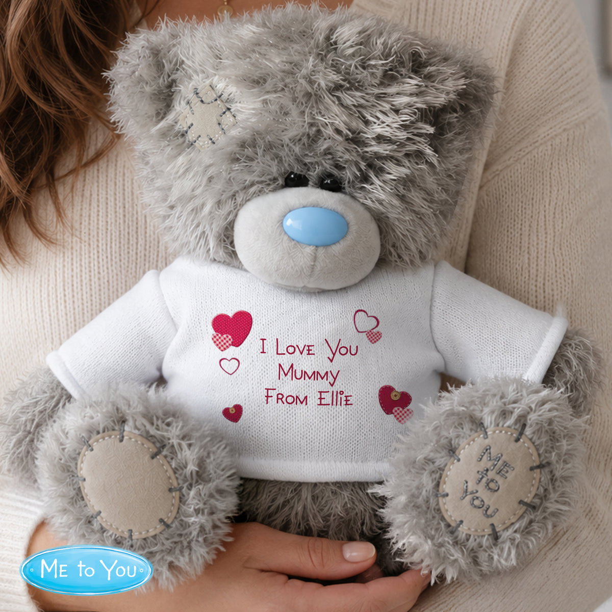 Grey teddy bear with a white shirt displaying 'I Love You Mummy From Ellie' held by a person.