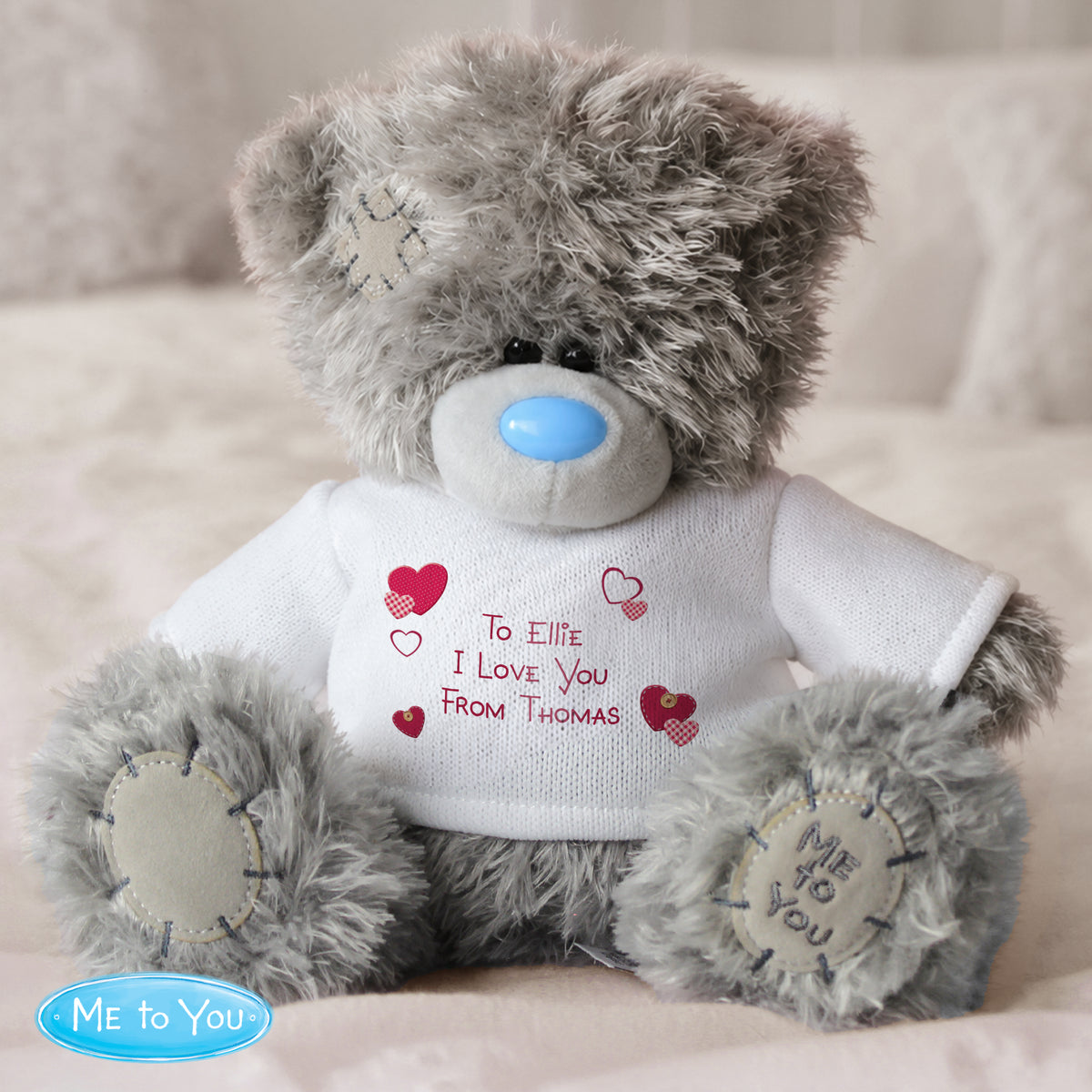 Personalised Grey teddy bear wearing a white shirt with text, sitting on a beige surface.