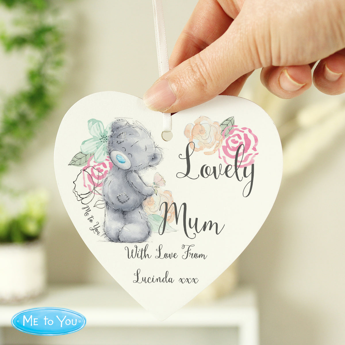 Heart-shaped decorative item with a teddy bear and floral design, held by a hand.