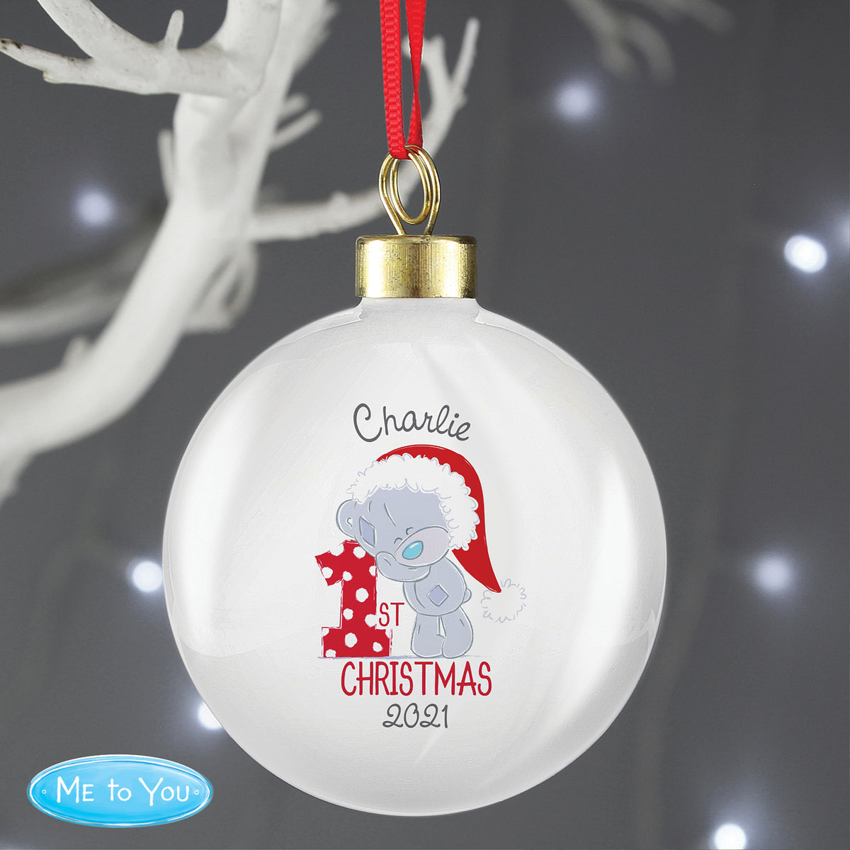 Christmas ornament with a teddy bear design and text, hanging against a dark background with blurred lights.