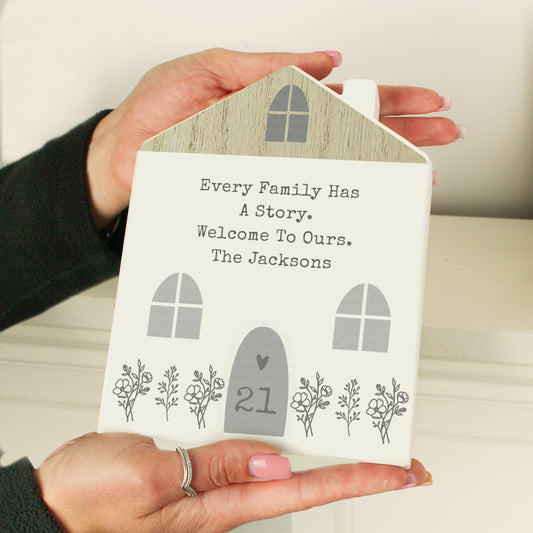 Personalised wooden house-shaped decoration held in hands, featuring the message "Every family has a story. Welcome to ours. The Jacksons" with floral detailing and customisable door number – a heartfelt home décor gift.