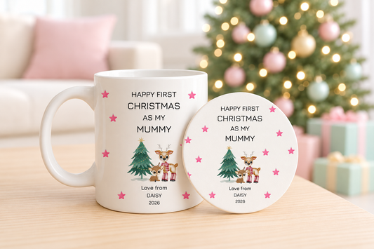 A white ceramic mug and matching round coaster set on a light wooden table, featuring a personalized Christmas design with pink stars, a small tree, two cartoon reindeer, and the text 'HAPPY FIRST CHRISTMAS AS MY MUMMY' and 'Love from DAISY 2026'. The background shows a softly blurred living room with a decorated Christmas tree and gifts.