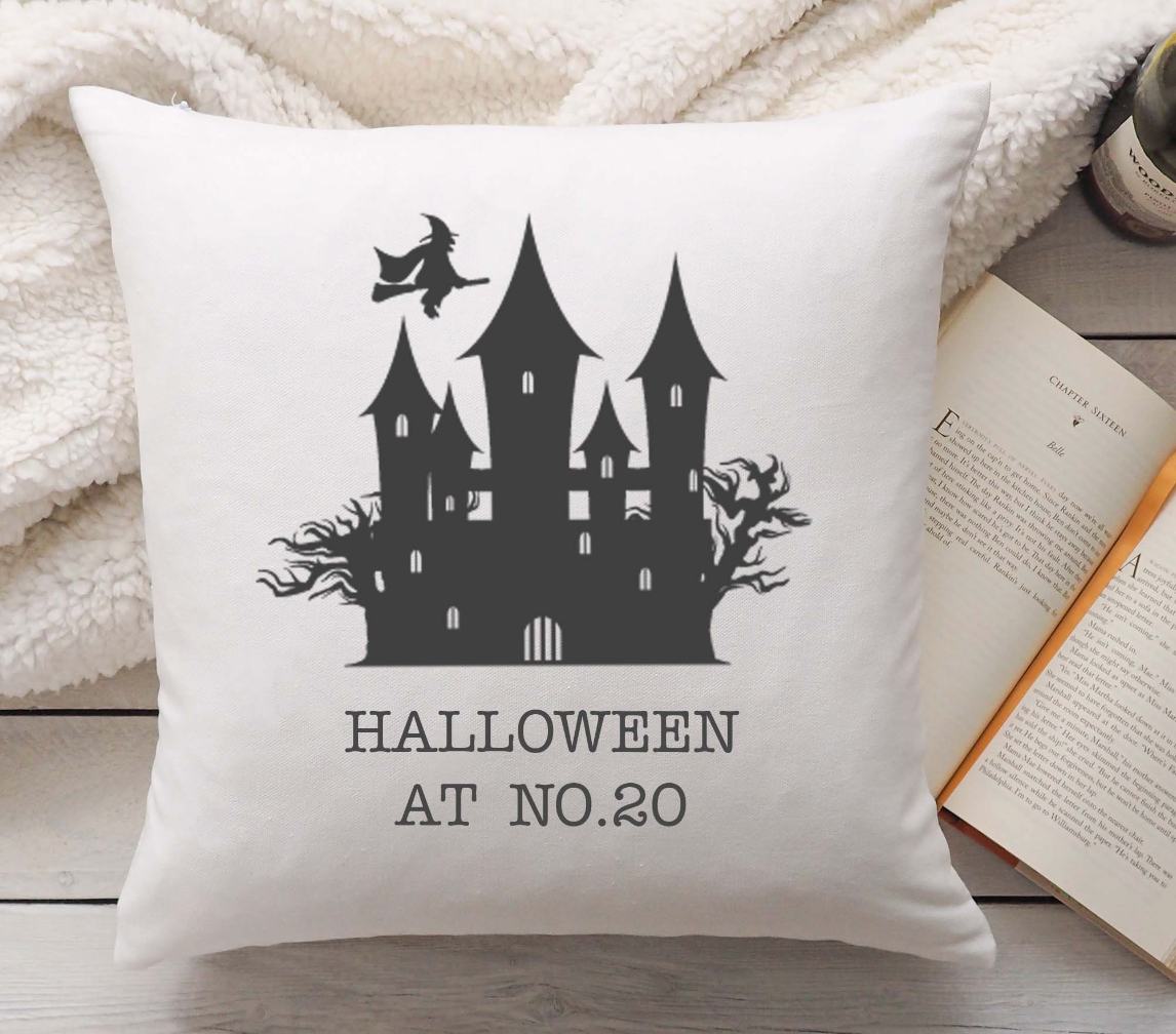 Linen pillow with black haunted house design and 'Halloween at No. 20' text, placed on a wooden surface with an open book.