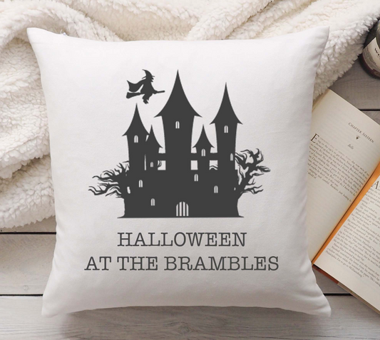 Linen pillow with Halloween-themed design featuring a castle and witch, placed on a wooden surface with a book and blanket.