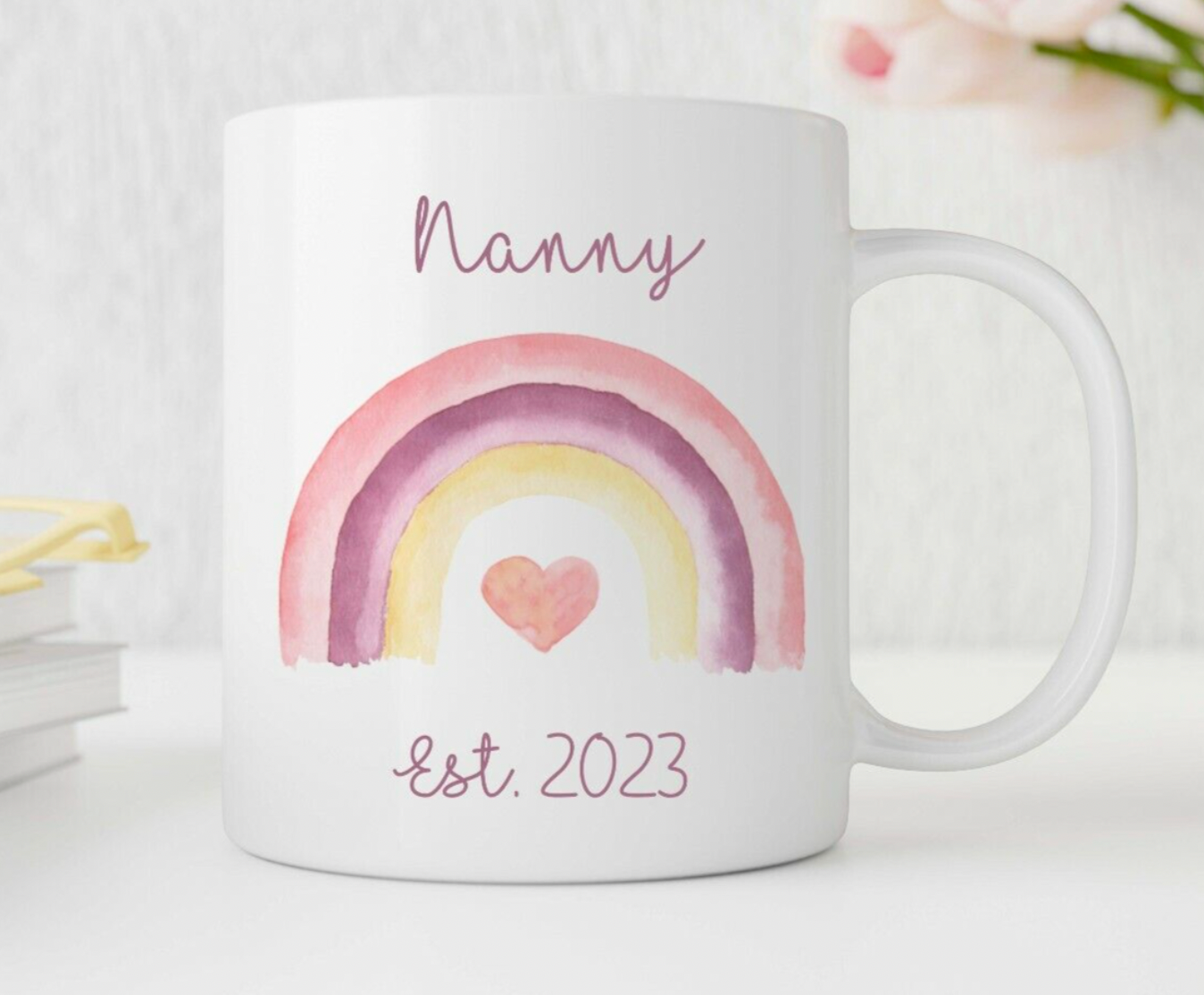 Personalised White mug with a rainbow and heart design, labeled 'Nanny Est. 2023', on a light background.