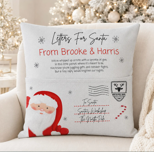 A square white throw pillow with a printed Christmas message for Santa, featuring a faux pocket design with postmarks, a Santa illustration, and a candy cane, displayed on a light-colored armchair.