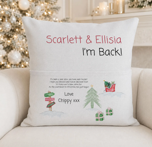 A personalized Christmas throw pillow on a beige sofa in front of a blurred Christmas tree and fireplace. The pillow is off-white with red and black personalized text reading 'Scarlett & Ellisia I'm Back!' and a small printed message with illustrations of a tree, gifts, and a dog in a sleigh.
