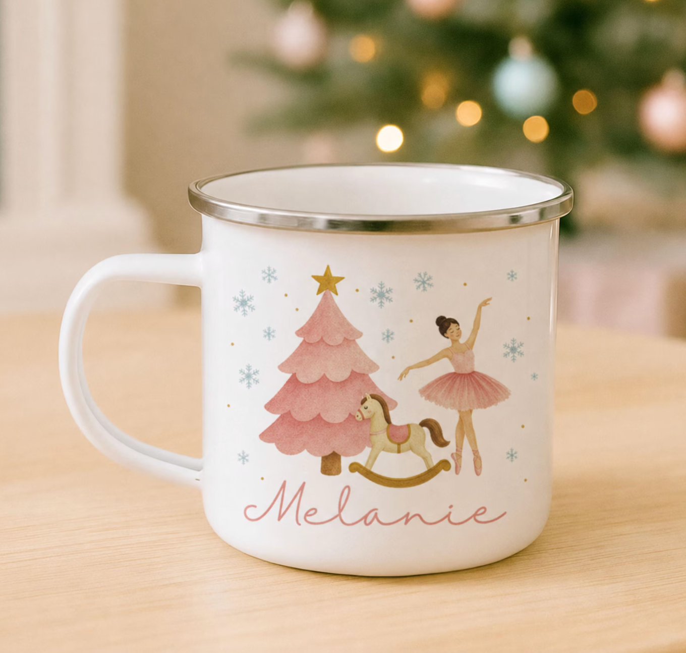 A white enamel-style mug with a silver rim sits on a light wood surface. The mug is printed with a festive illustration featuring a ballerina, a pink Christmas tree, a rocking horse, snowflakes, and the name 'Melanie' in pink script.