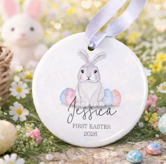 Round ceramic ornament with a bunny design and 'First Easter 2026' text, hanging on a blurred Easter-themed background.