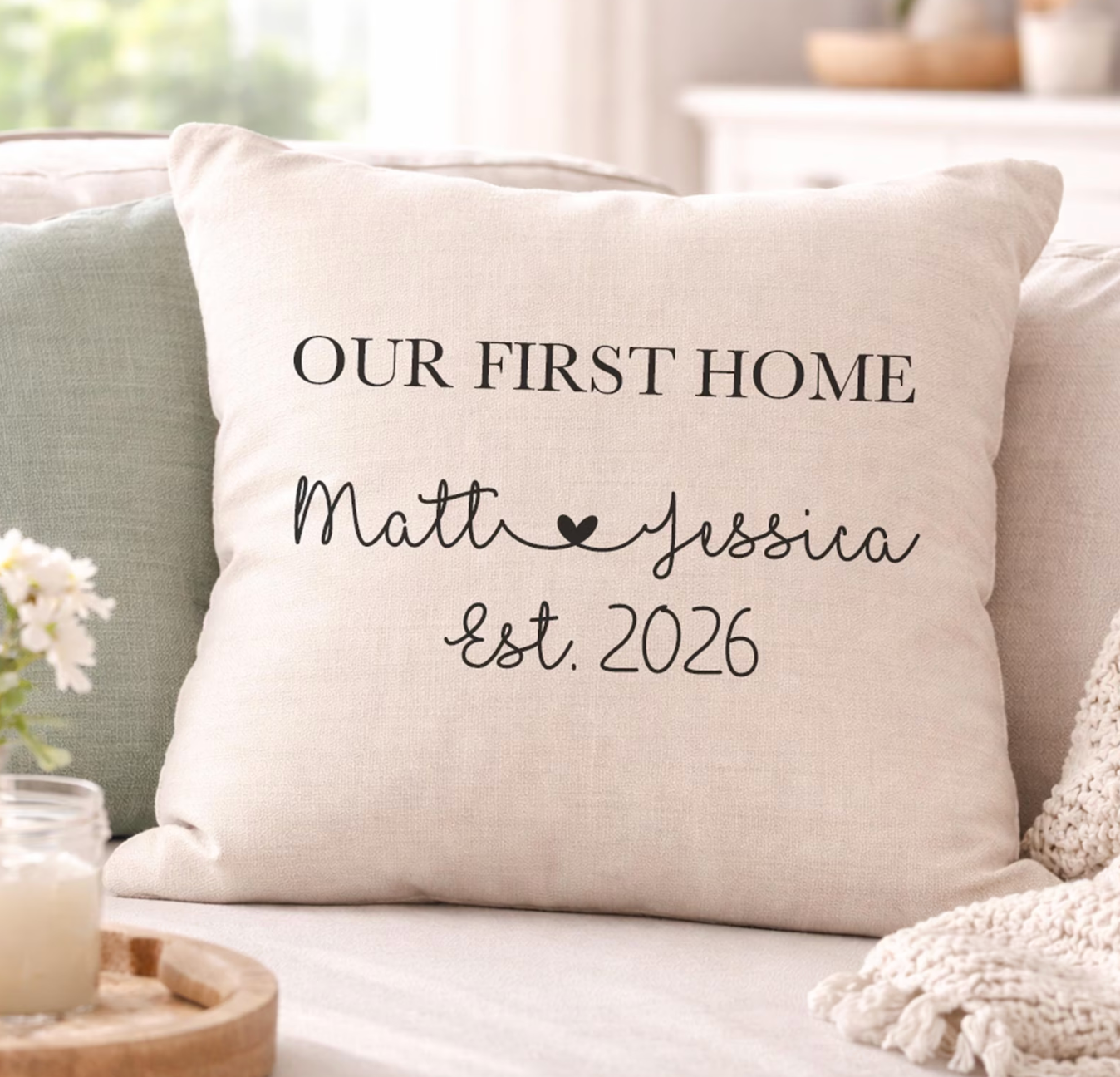A personalised throw pillow on a sofa with the text 'OUR FIRST HOME Matt & Jessica Est. 2026' printed in black.