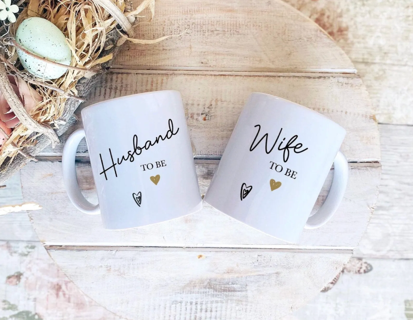 Two white ceramic mugs with black text reading 'Husband TO BE' and 'Wife TO BE', decorated with small gold hearts, shown on a rustic wooden surface with a decorative nest and eggs in the corner.