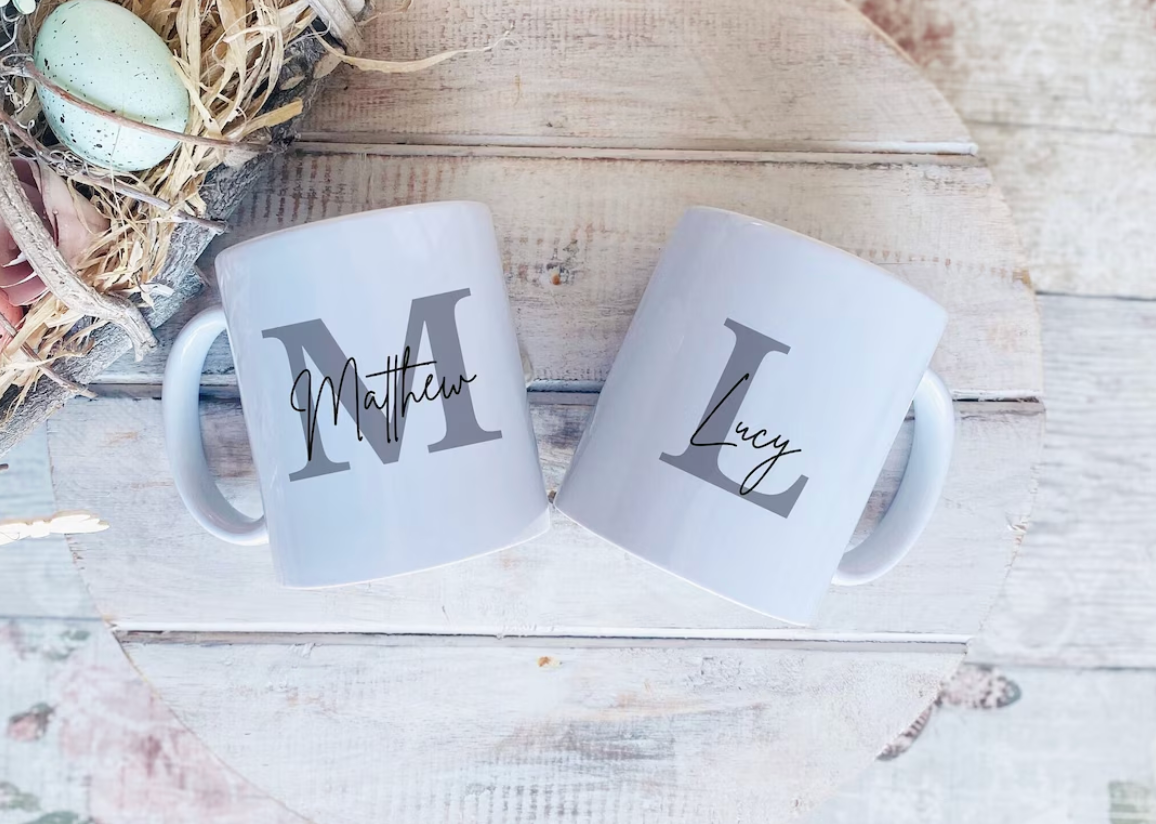 Two white ceramic mugs with custom monogram names 'Matthew' and 'Lucy' printed in gray, displayed on a rustic white wooden surface next to a decorative Easter egg nest.