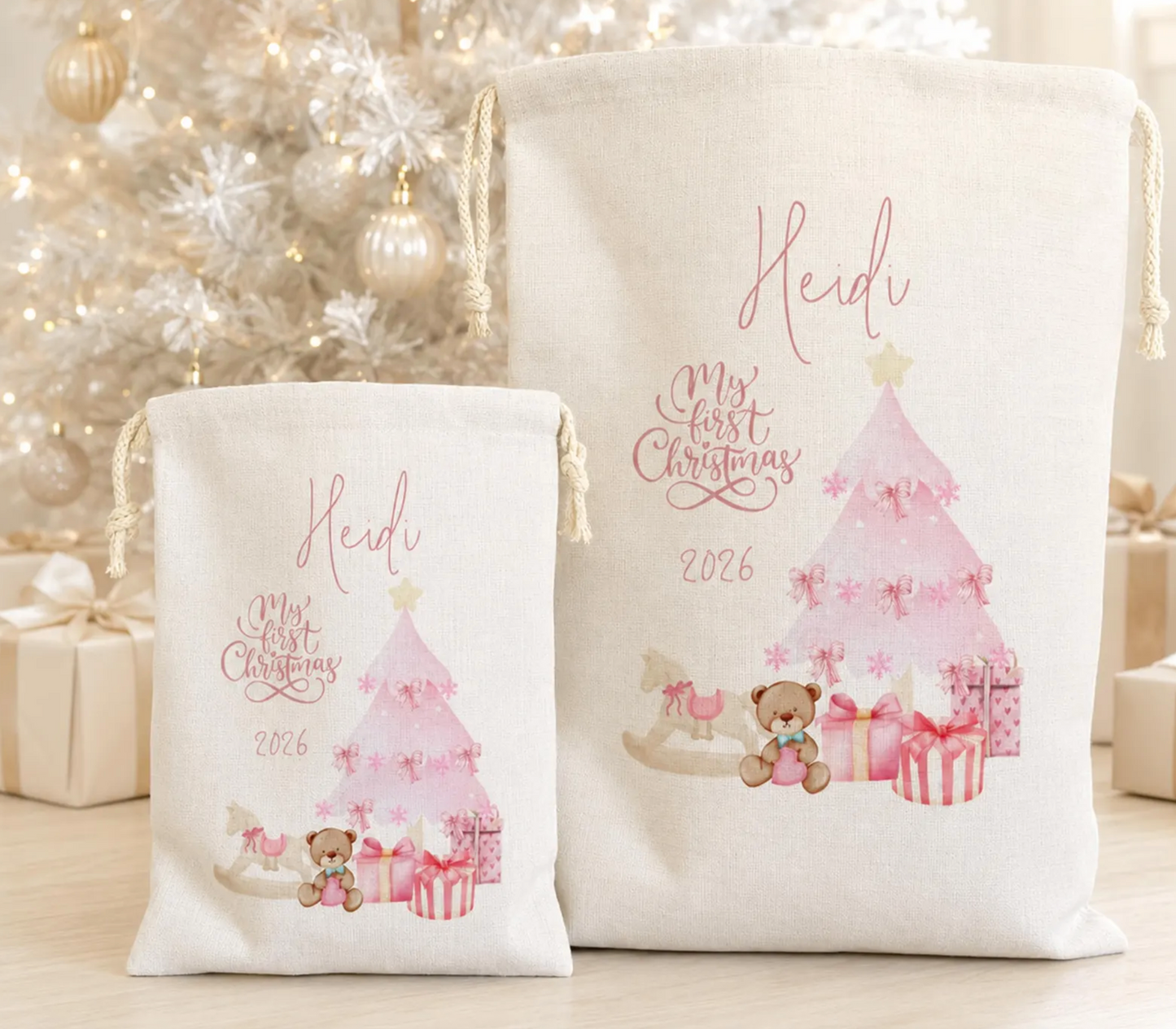 Two personalized cream canvas drawstring gift bags displayed in front of a softly blurred white Christmas tree. The larger bag features a pink watercolor Christmas tree, presents, a teddy bear, and the text 'Heidi', 'My First Christmas', and '2026'.